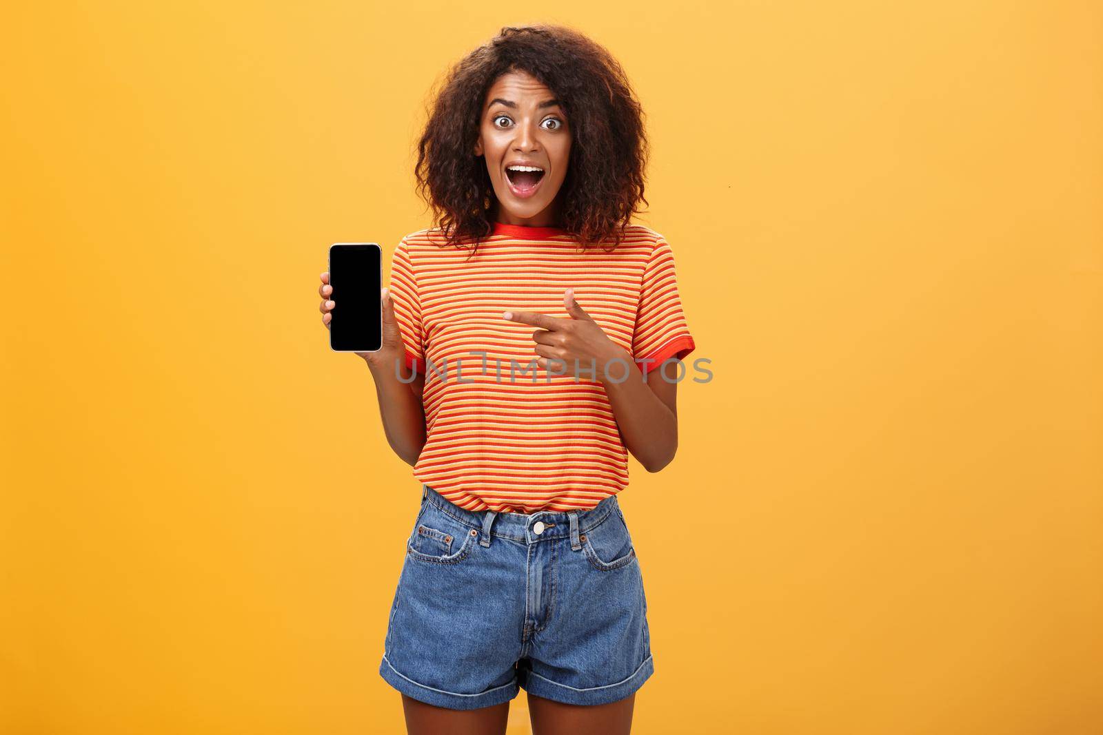 Impressed fascinated stylish slim african american curly-haired woman in striped t-shirt dropping jaw from amazement holding awesome smartphone pointing at device screen over orange wall. Lifestyle.