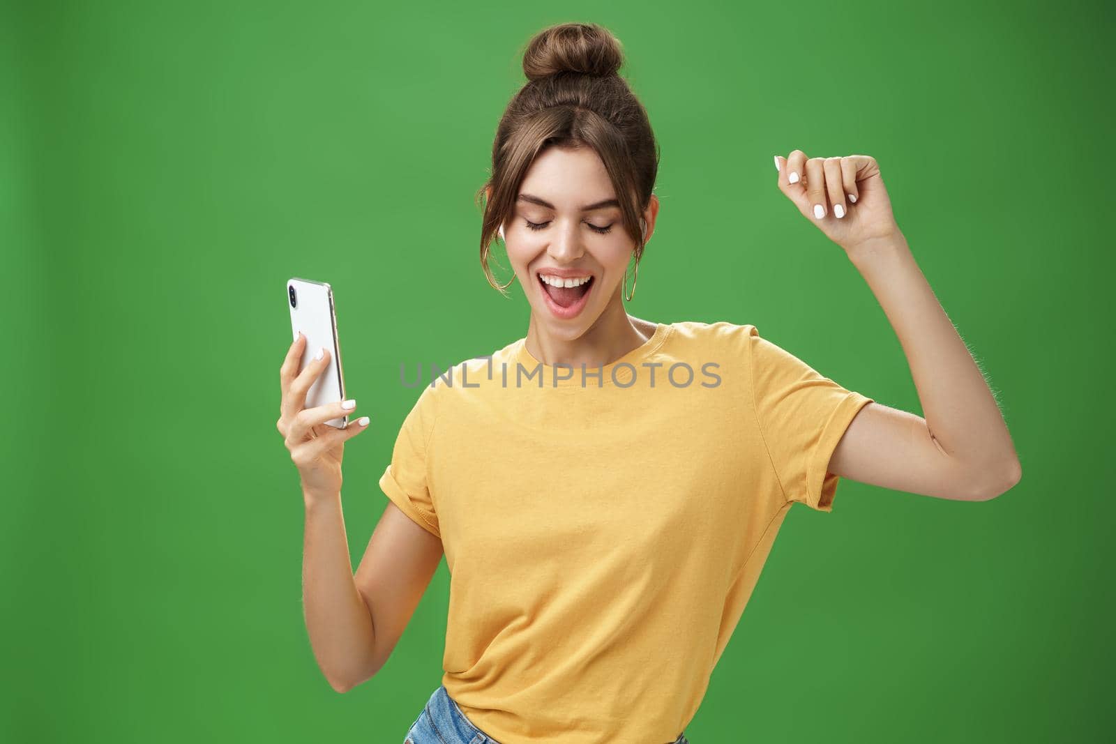 Cool and stylish happy woman with tattoo and gap teeth in yellow t-shirt dancing joyfully listening music in wireless earphones holding smartphone closing eyes and smiling from delight over green wall by Benzoix