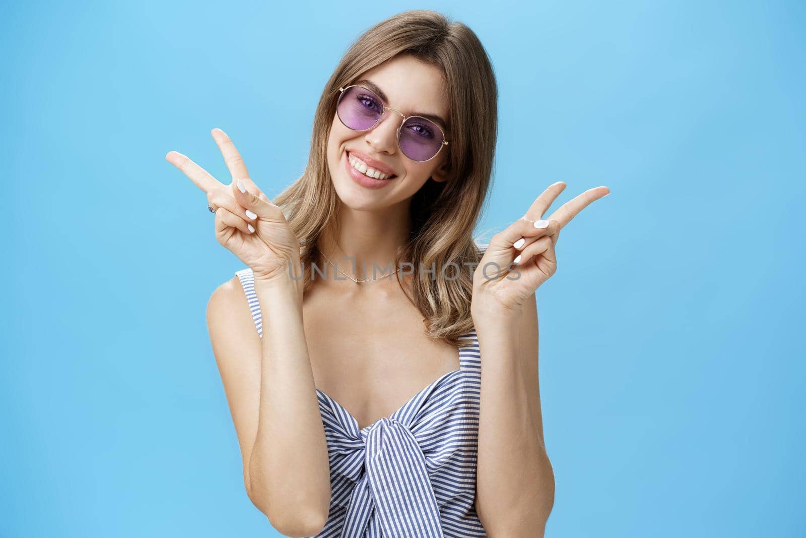 Waist-up shot of cute self-assured feminine girlfriend in trendy top showing peace gestures near shoulders tilting head amused smiling broadly showing off gapped teeth posing over blue wall by Benzoix