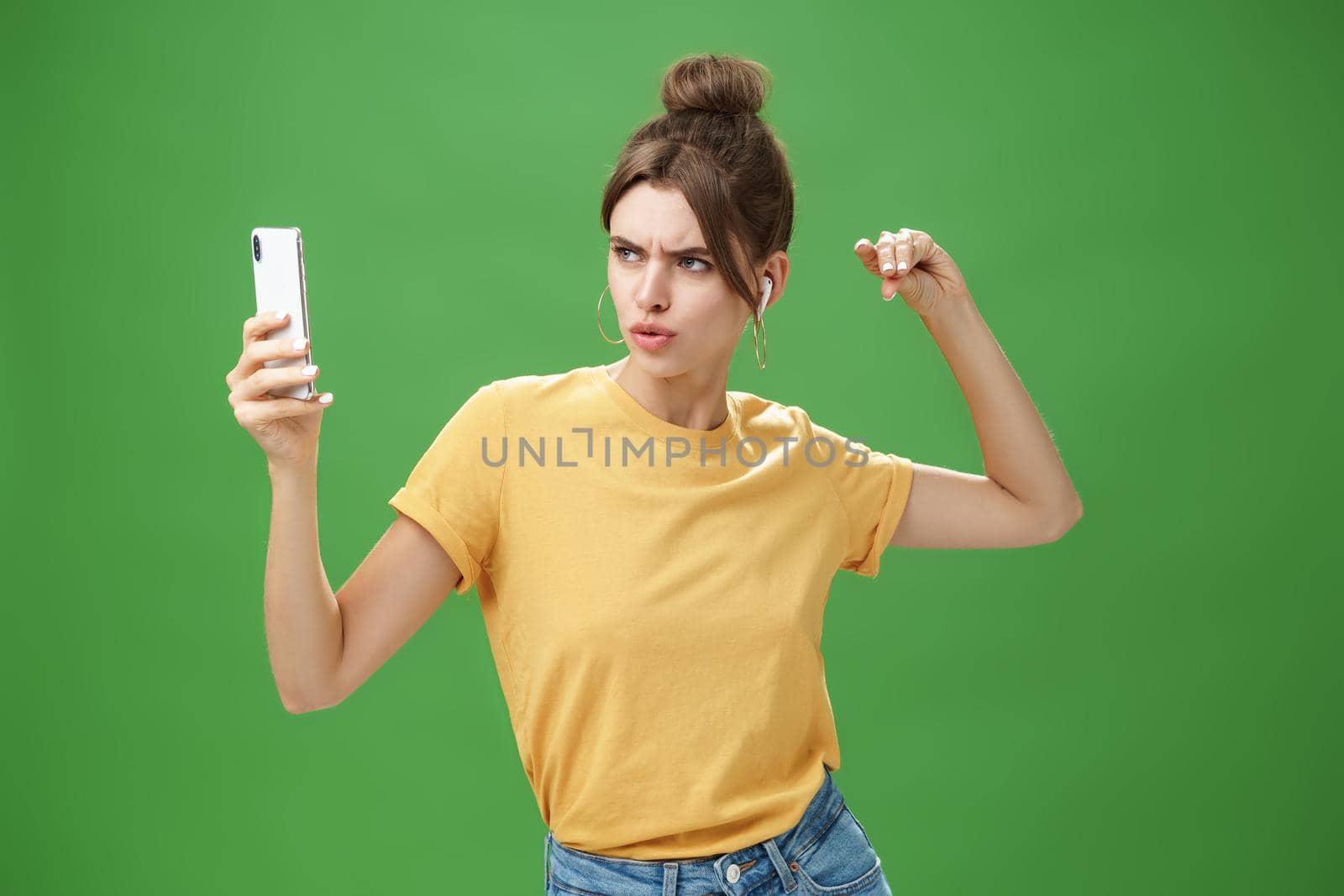 Woman making serious look to take selfie while listening music in wireless earphones posing and staring at smartphone screen daring and aggressive, liking post photos online over green background. Technology and lifestyle concept