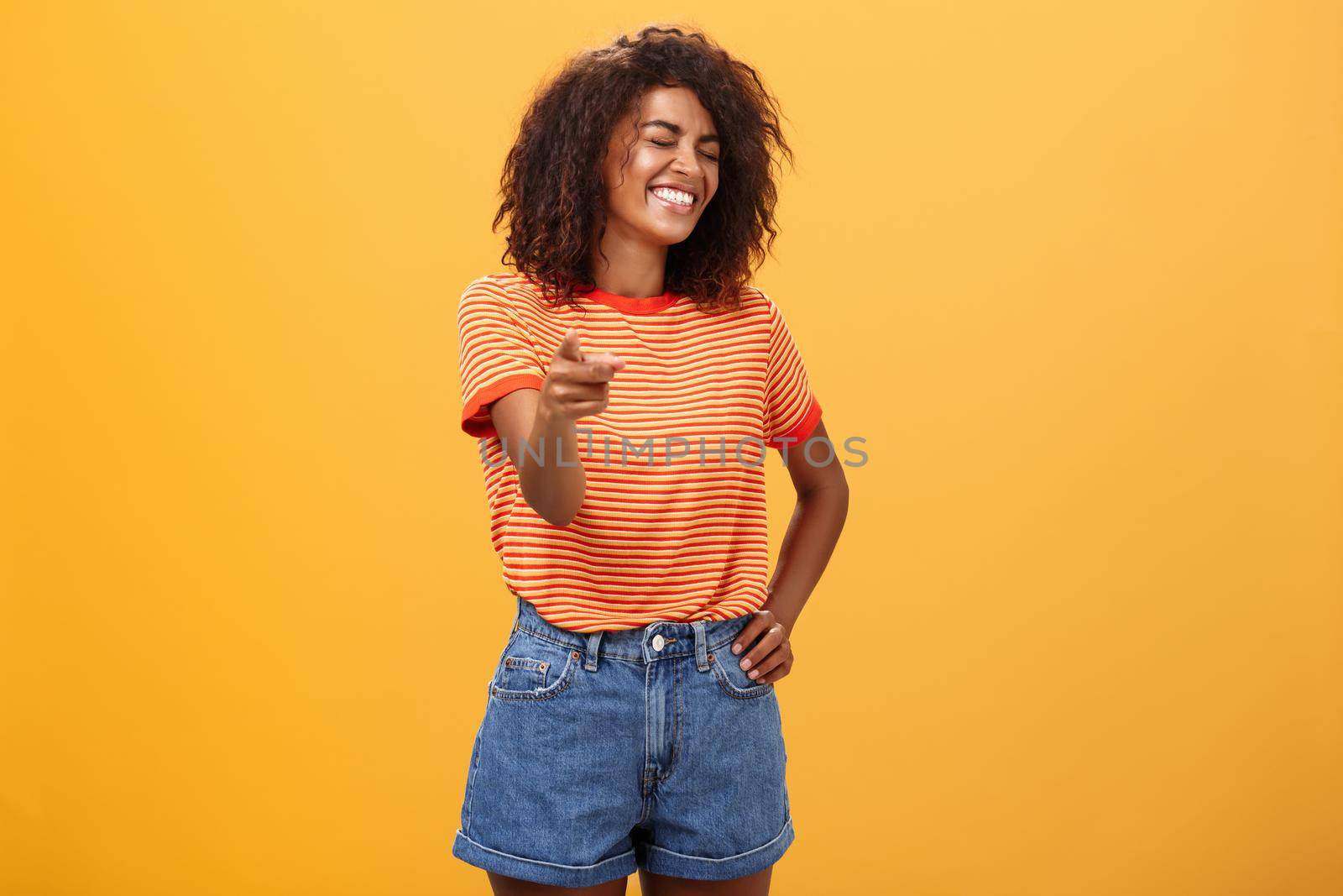 Girl pointing at funny and hilarious friend approving good joke. Amused and entertained attractive dark-skinned female with afro hairstyle laughing with closed eyes indicating at camera joyfully.