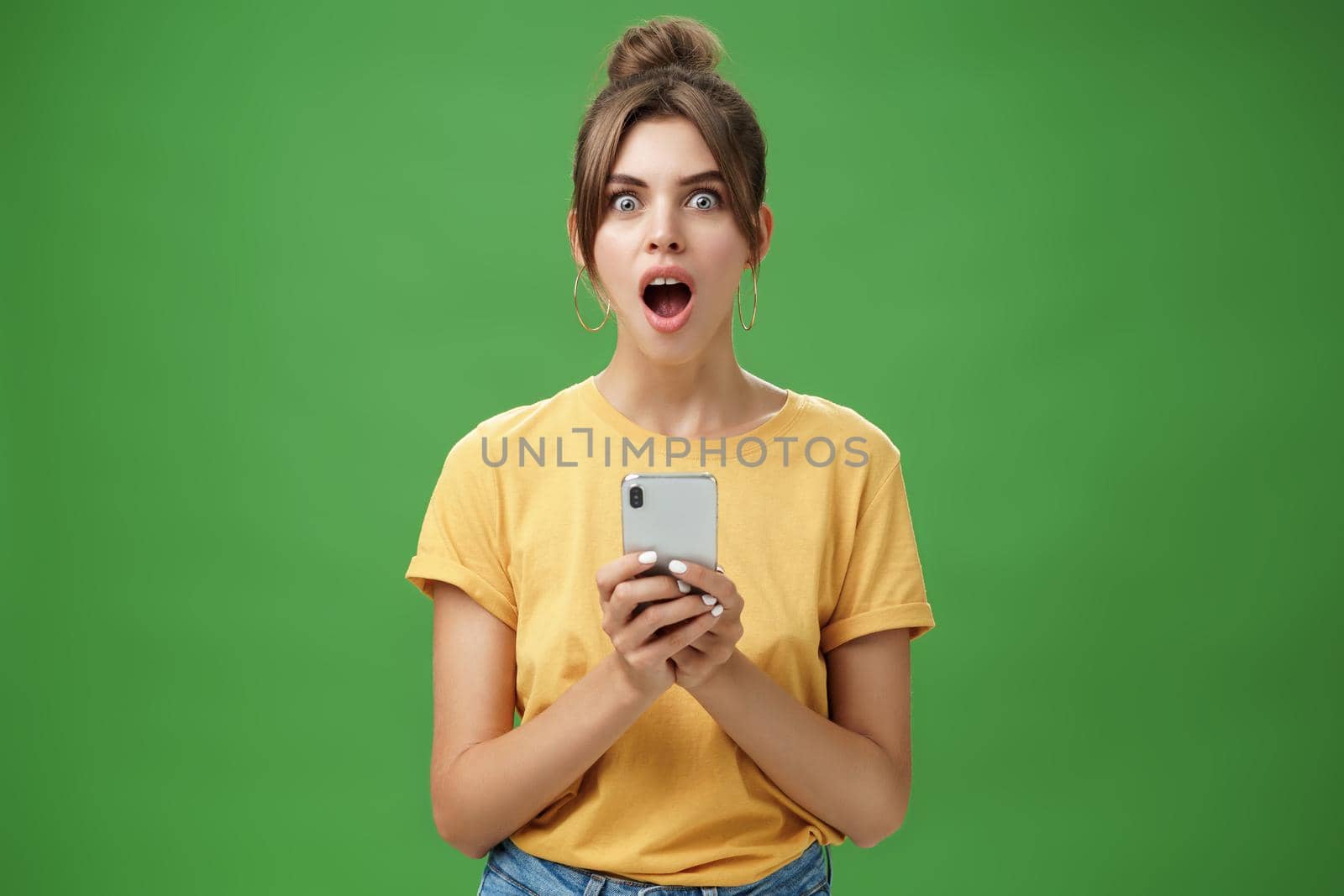 Portrait of shocked speechless and impressed beautiful white girl with combed hair in yellow t-shirt holding smartphone, dropping jaw from excitement reacting to cool app over green background.