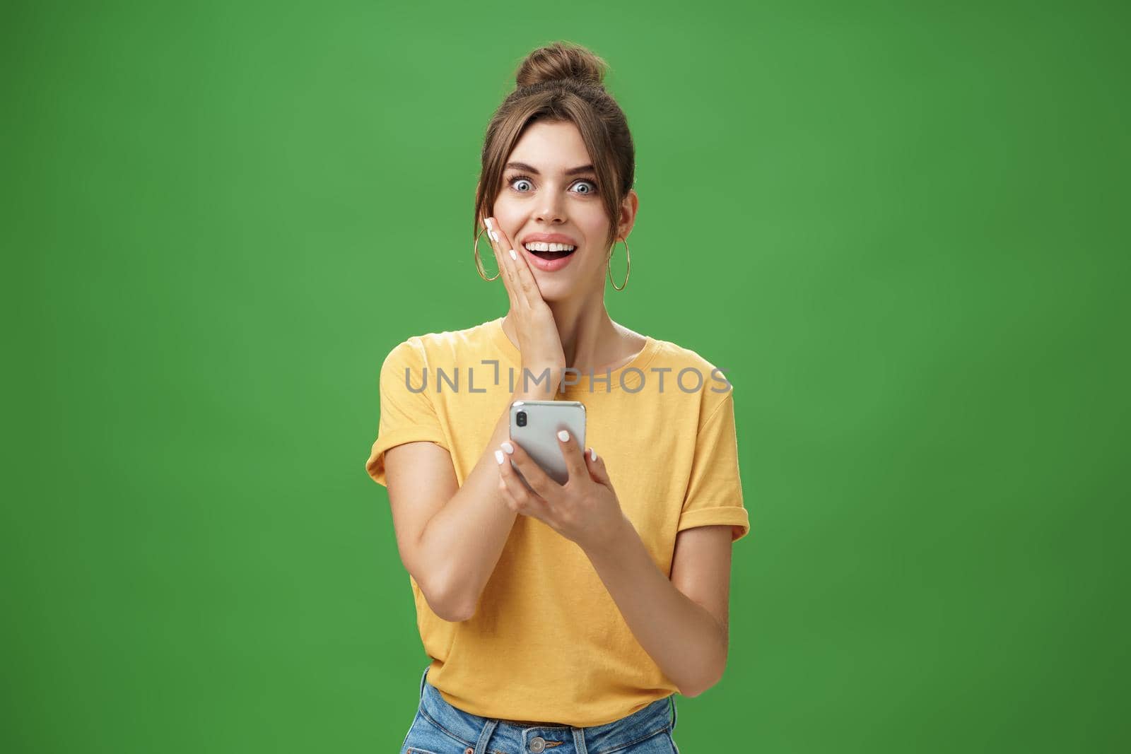 Surprised touched and impressed beautiful european girl with diastema smiling broadly touching cheek from amazement and joy holding smartphone looking happy at camera after reading cool news by Benzoix