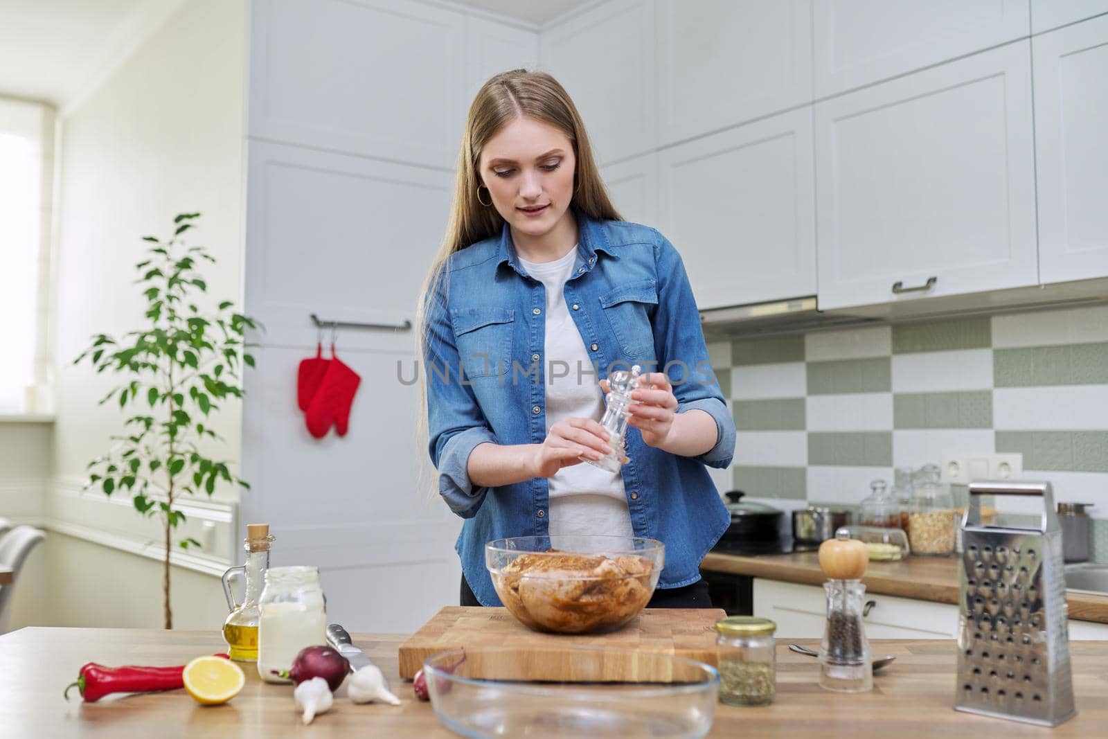 Young woman cooking chicken, marinating with spices black pepper salt, at home in kitchen by VH-studio