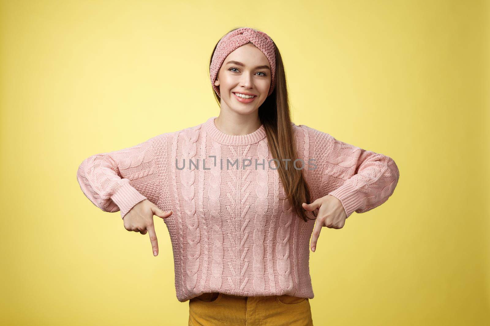 Tender young 20s european girl in sweater smiling broadly finally take-off braces pointing down promoting skillful dentist professional. Female student standing happy positive over yellow wall by Benzoix