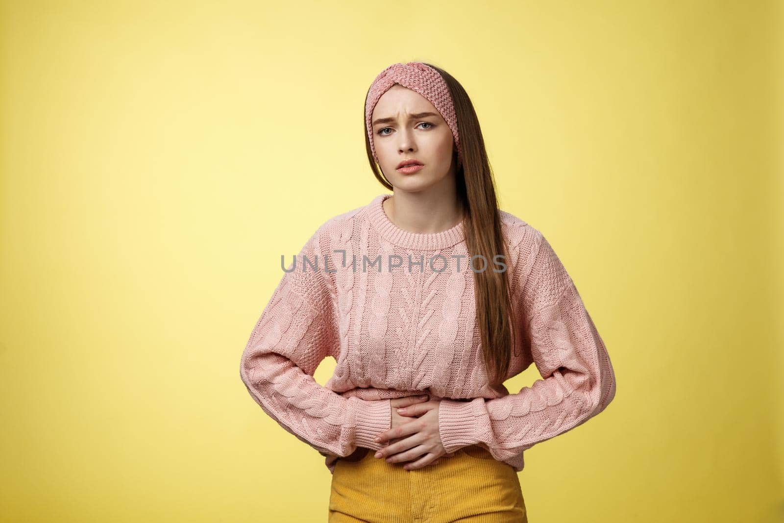 Girl suffering pain in stomach, having painful periods, feeling unwell touching belly grimacing displeased and troubled, having cramps, stooping upset of stomachahe against yellow background.