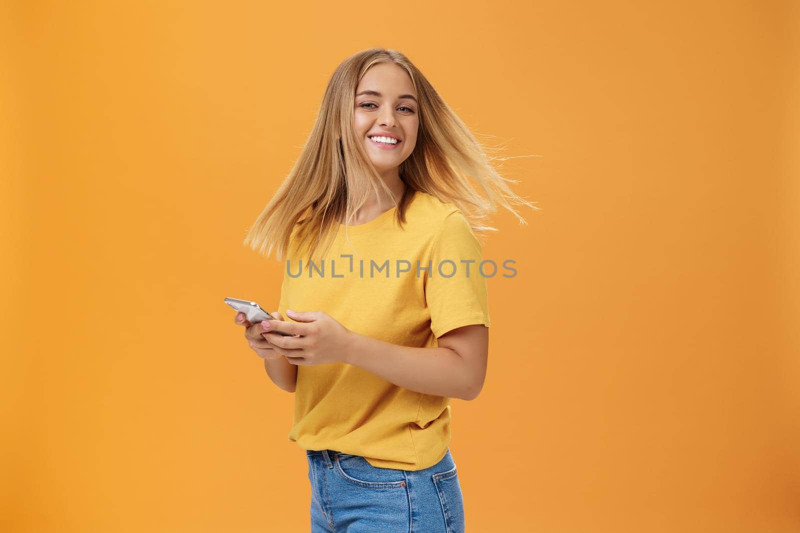 Girl feeling happy receiving message from him. Portrait of carefree charming woman with new haircut waving head standing half-turned, turning to camera with pleased cute smile holding smartphone by Benzoix