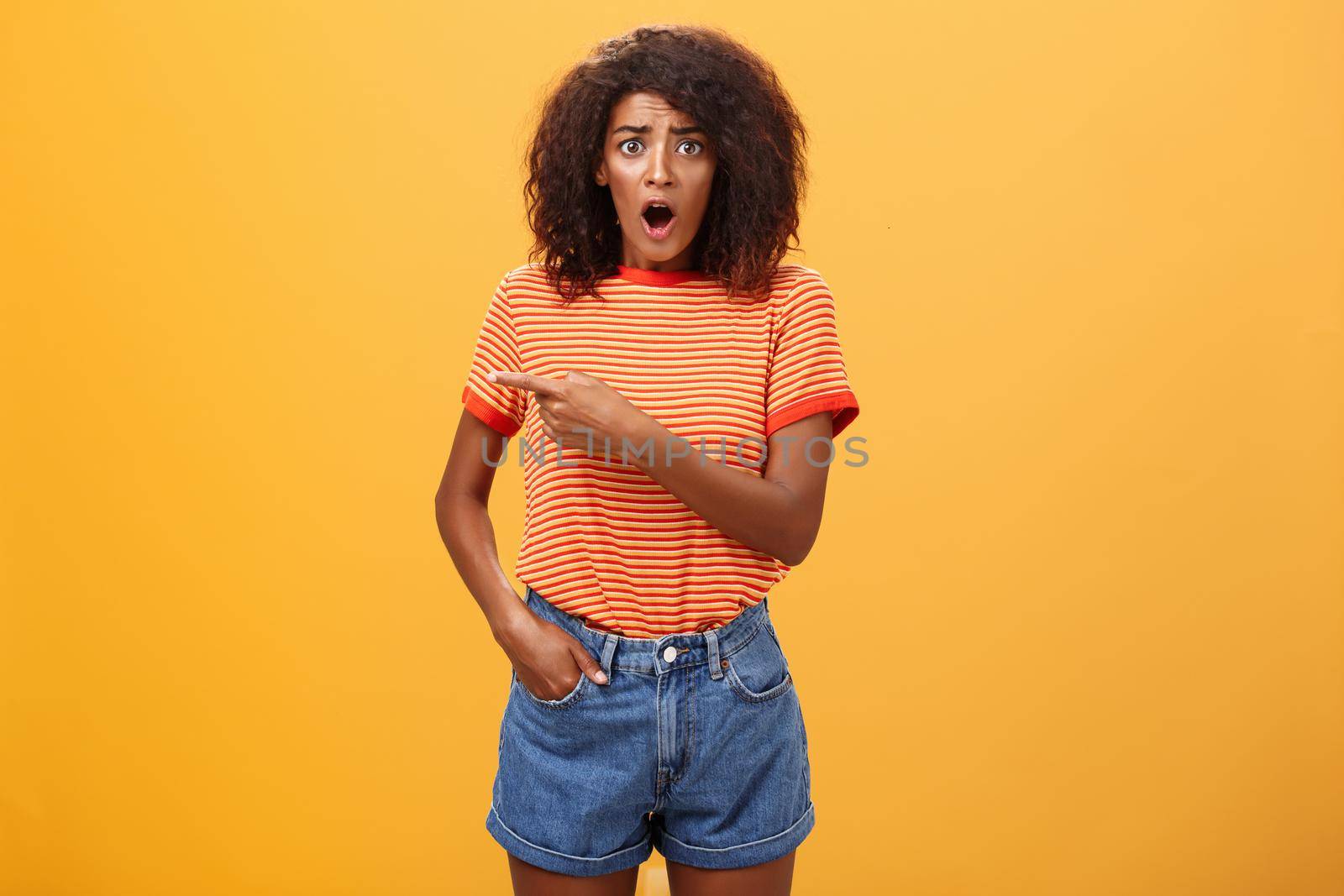 Studio shot of offended shocked stylish woman being robbed fasping and staring nervously begging stranger help out pointing left concerned and worried frowning, posing displeased over orange wall.