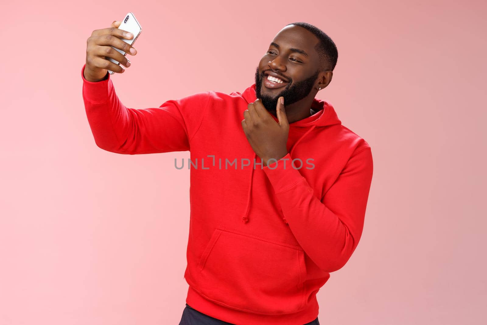 Cheeky cool african american confident bearded guy in red hoodie taking selfie update date app profile photo holding smartphone raised touching face sassy smiling look phone display flirty.