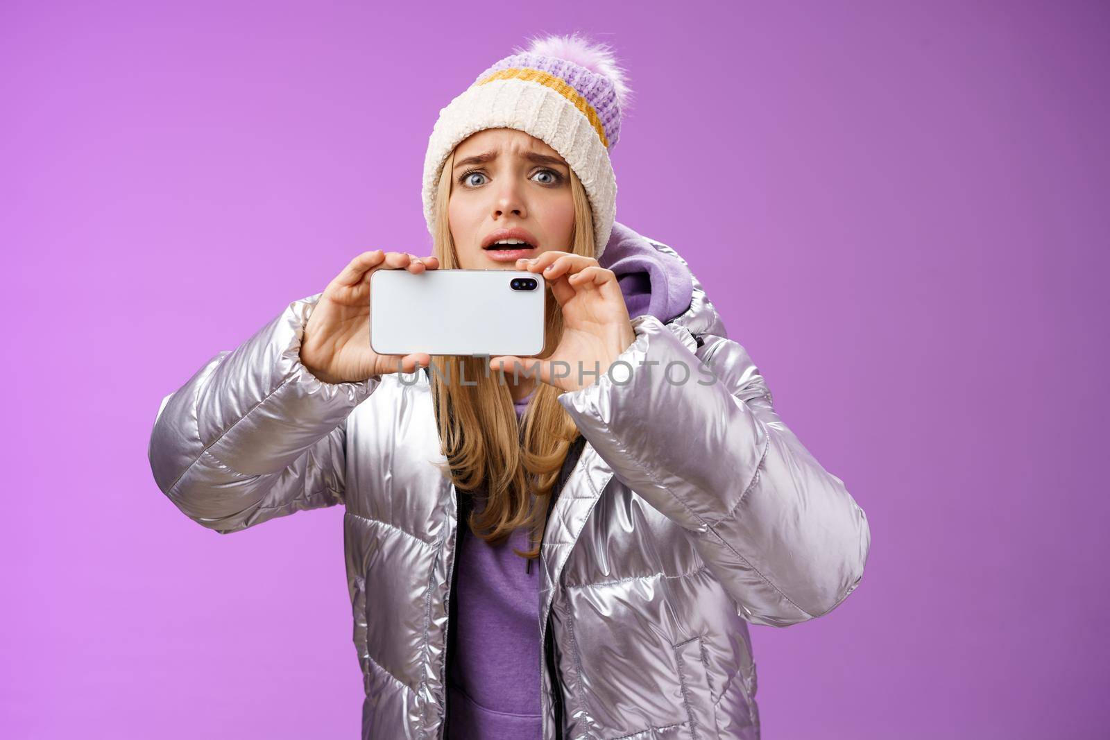 Intense worried blond girl holding mobile phone horizontal recording video capture moment share friends internet blog using smartphone camera shoot photograph standing serious purple background.