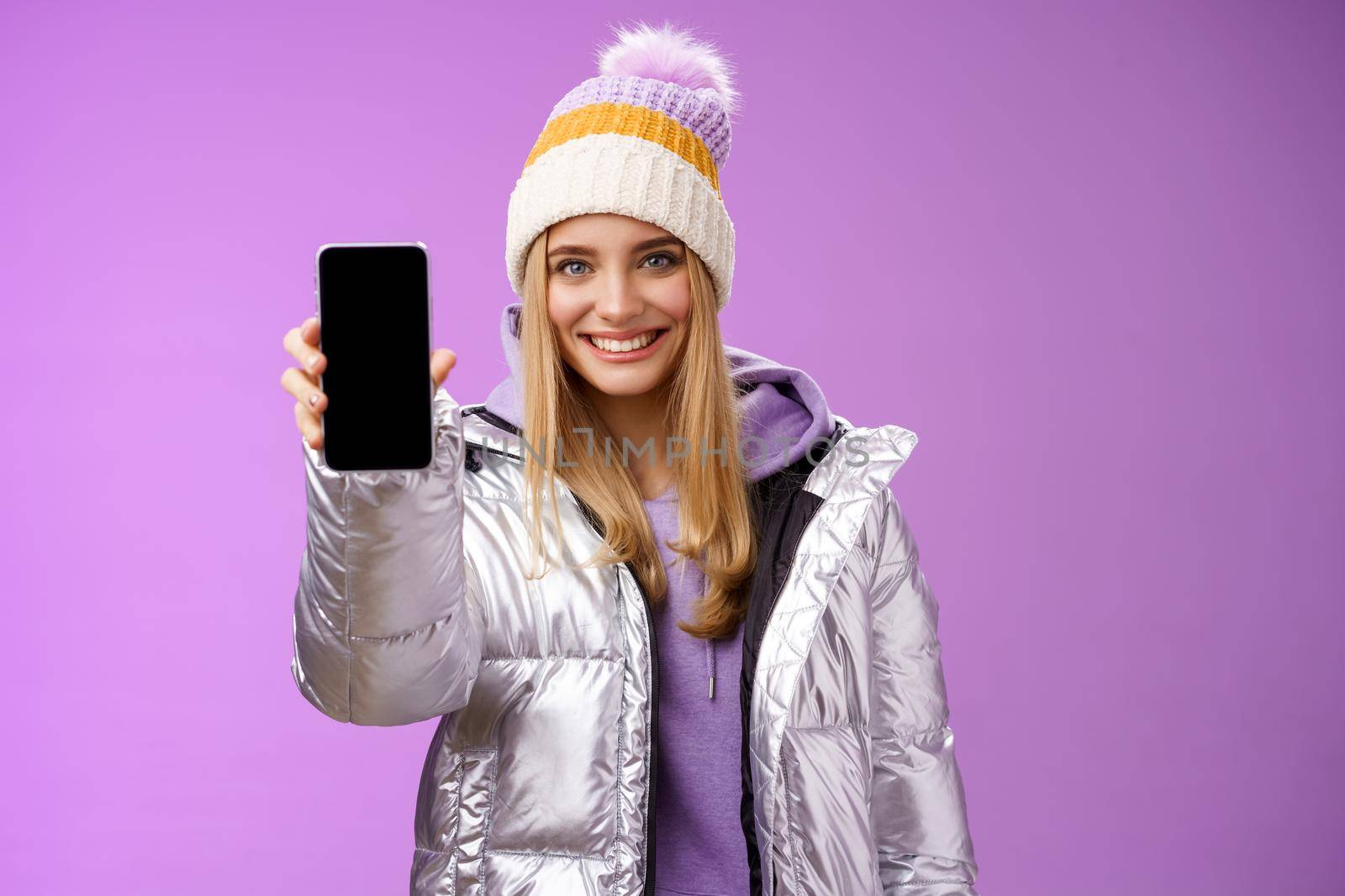 Lifestyle. Friendly cheerful confident blond girl in silver stylish winter jacket hat extend arm showing smartphone display advertising awesome new device app smiling self-assured recommend use mobile phone.