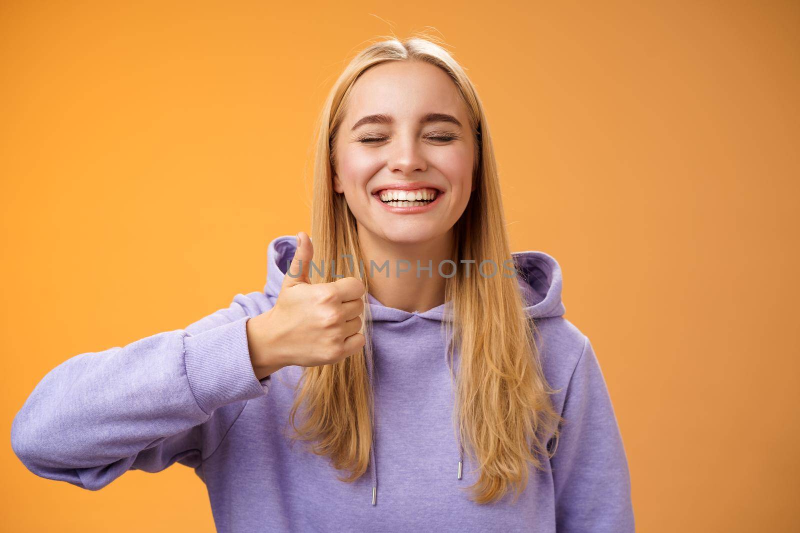 Amused joyful optimistic young excited blond european female model in hoodie smiling broadly close eyes having fun show approval gesture thumbs-up like awesome trip vacation idea, orange background.