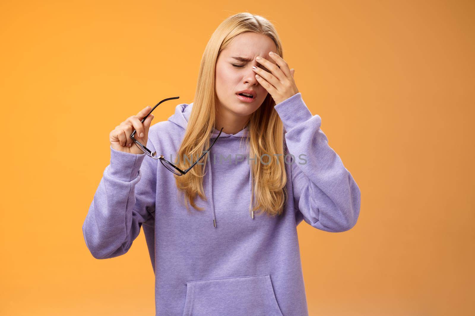 Tired dizzy cute blond girl taking off glasses touching forehead feel sick unwell suffering headache painful migraine standing bothered uncomfortable orange background in hoodie.