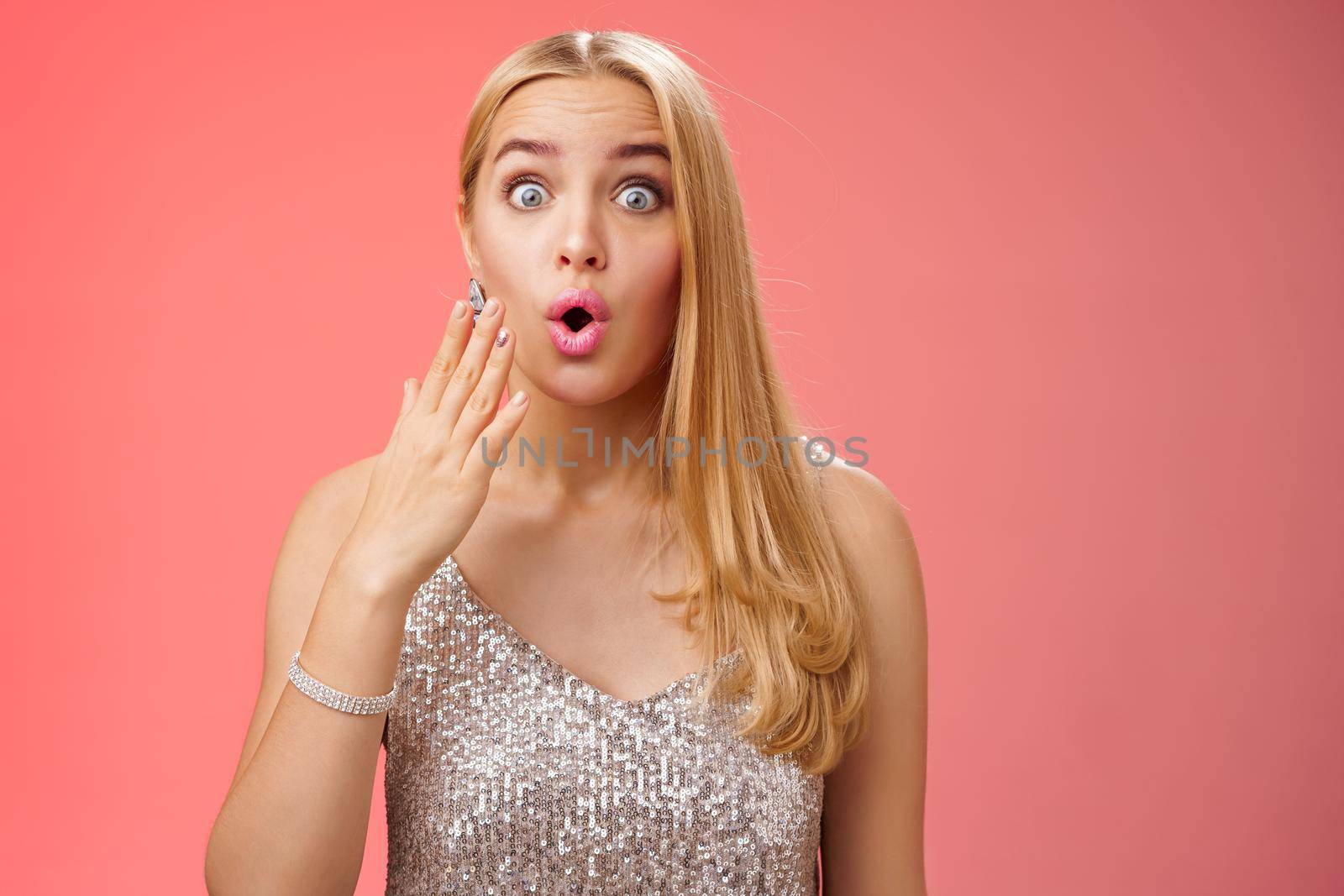 Amused wondered good-looking elegant blonde in silver stylish dress pop eyes surprised folding lips wow cover mouth palm amazement learn incredible rumor gossip with friend, red background.