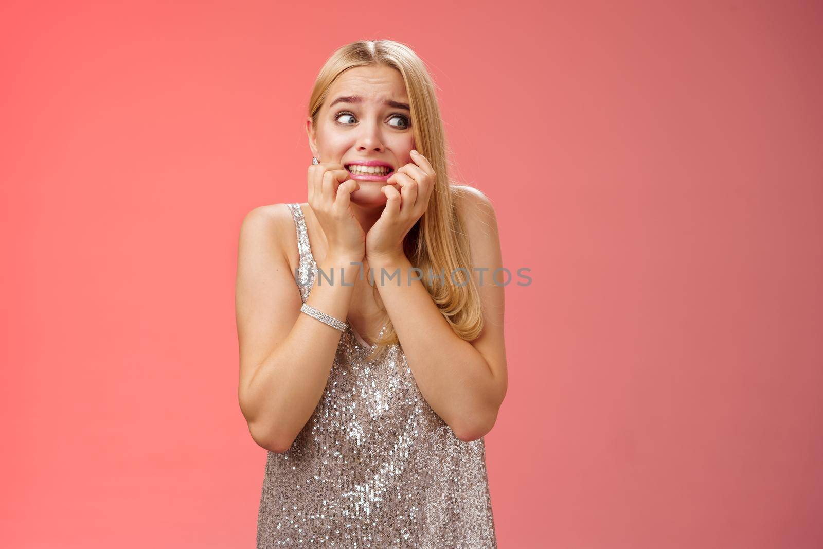 Frightened insecure timid young blond woman afraid feel scared trembling fear stooping biting nails pop eyes right standing horrified red background wear silver evening dress terrified by Benzoix