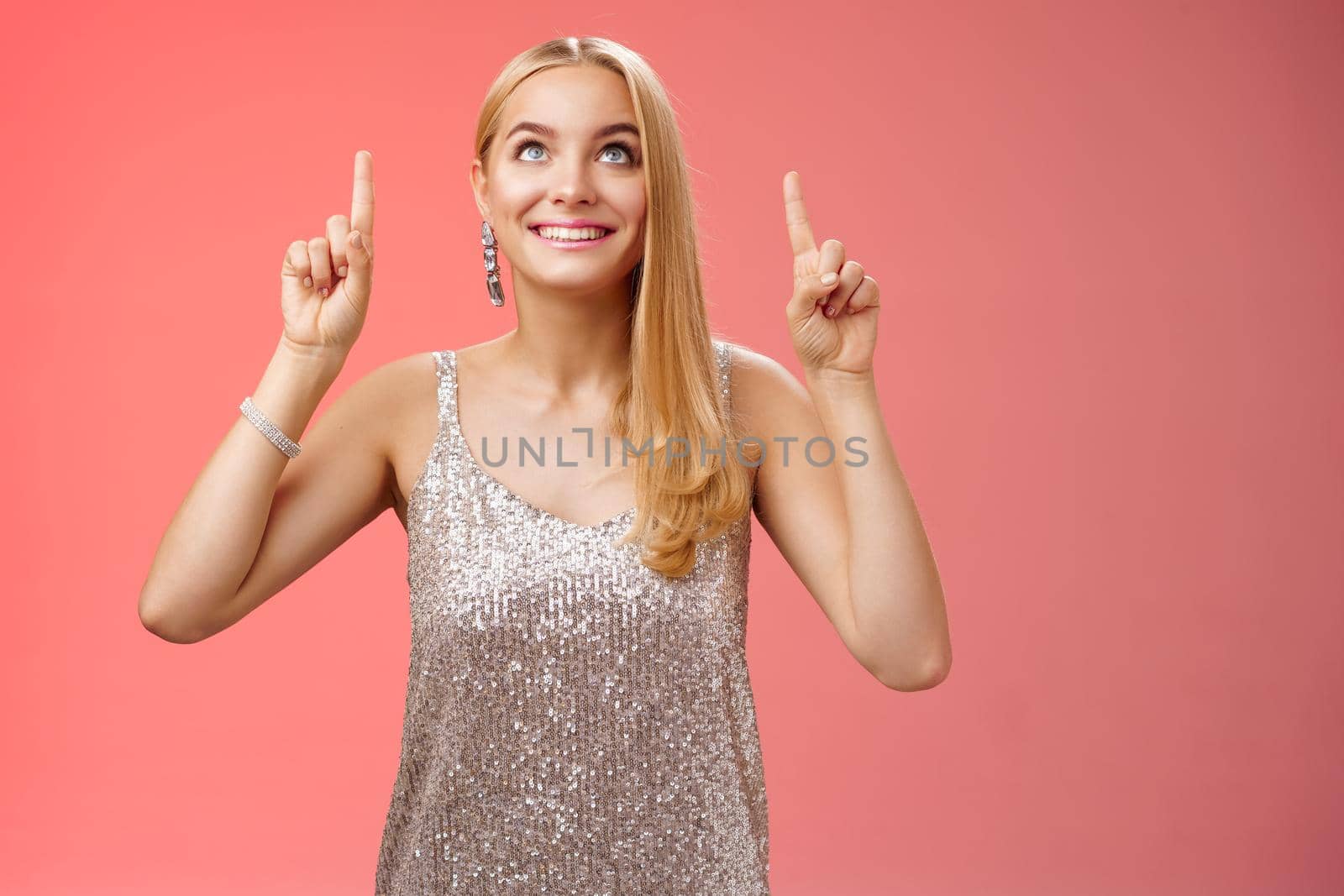 Amused gorgeous tender elegant european woman blond long hairstyle in silver glittering evening dress look pointing up curious gaze excitement see desired thing, standing red background joyful.