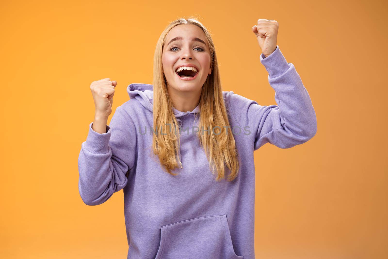 Happy cheerful young supportive woman cheering sister win smiling dedicated fun hope favorite team score goal raising clenched fists yelling happily celebrating triumphing victory, orange background.