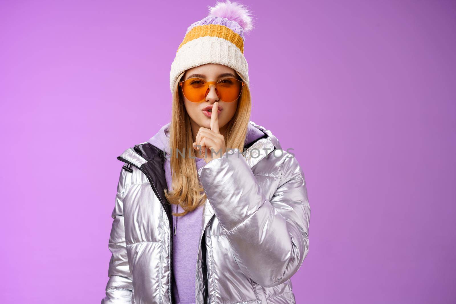 Sassy flirty gorgeous woman enjoying vacation snowy mountain resort wear winter hat silver stylish jacket sunglasses asking promise not tell secret show shush shhh gesture index finger seal lips by Benzoix
