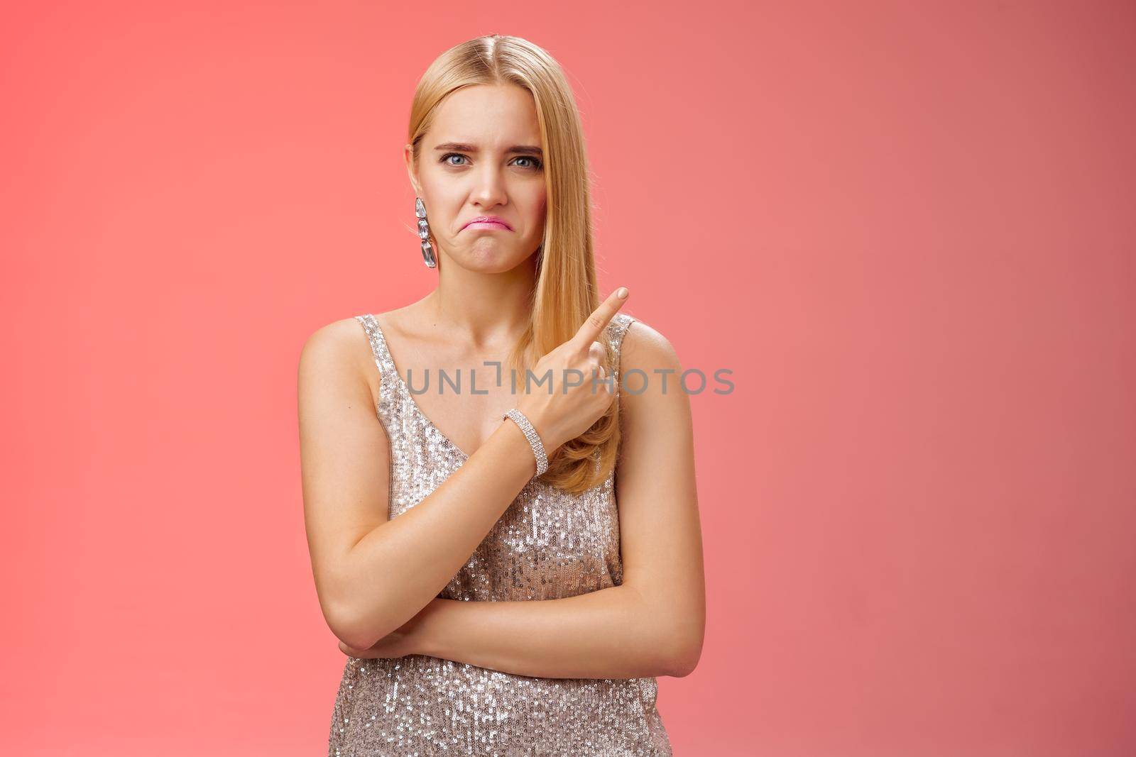 Jealous displeased angry young revengeful blond ex-girlfriend in luxurious silver shiny dress frowning pouting pointing upper right corner displeased pissed standing fed up upset red background by Benzoix