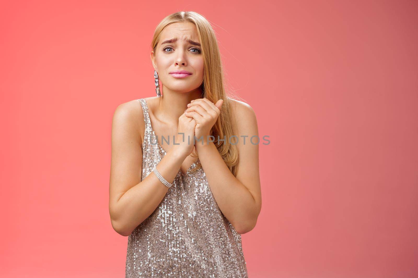 Sad pity charming blond european hospitable woman feel empathy touching upsetting story press palms together heart frowning crying sorry for friend, standing in silver dress red background.