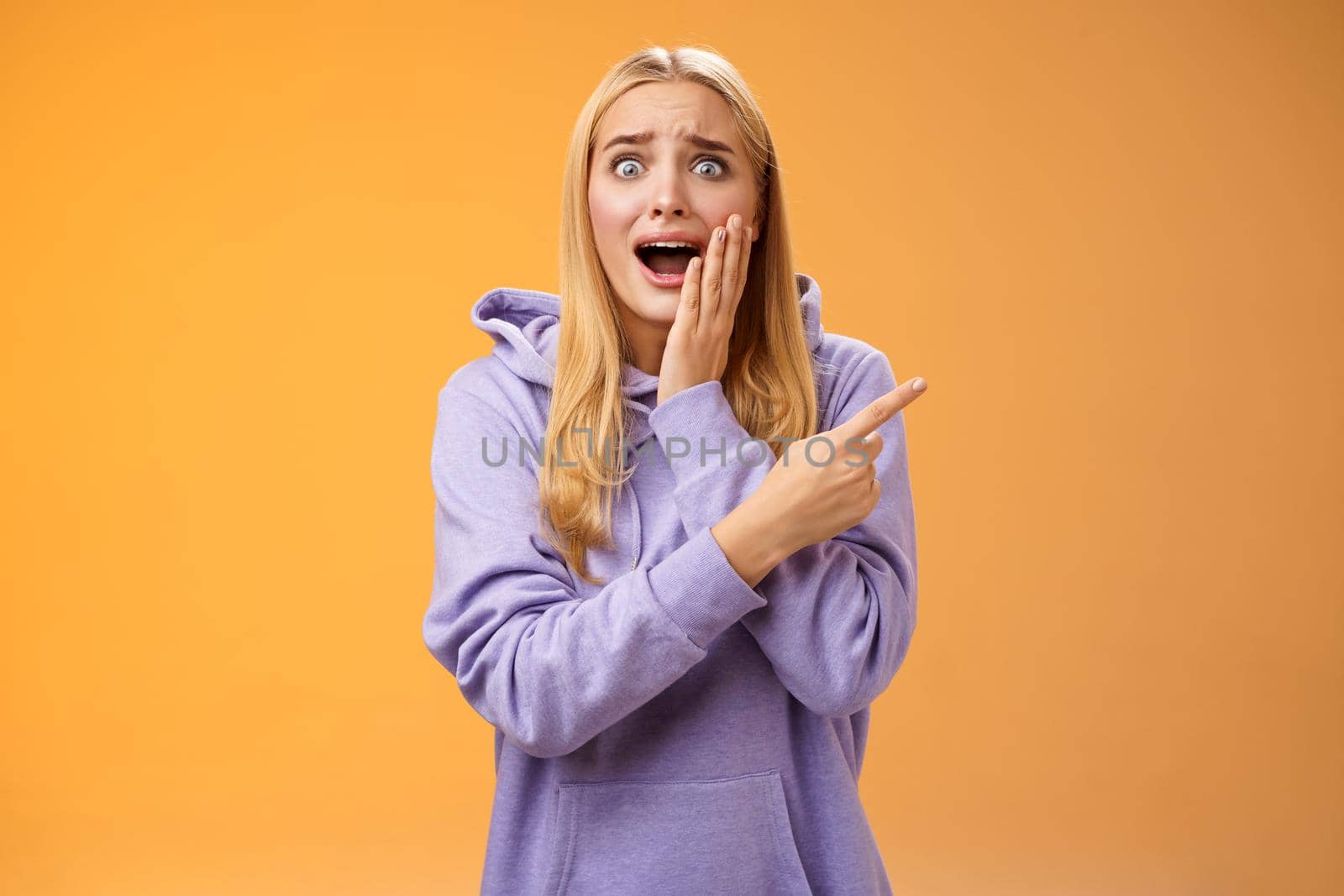 Amazed excited overwhelmed young blond girl in hoodie press palm cheek screaming astonished pointing right seeing famous super star cannot hold joyful positive emotions, orange background.