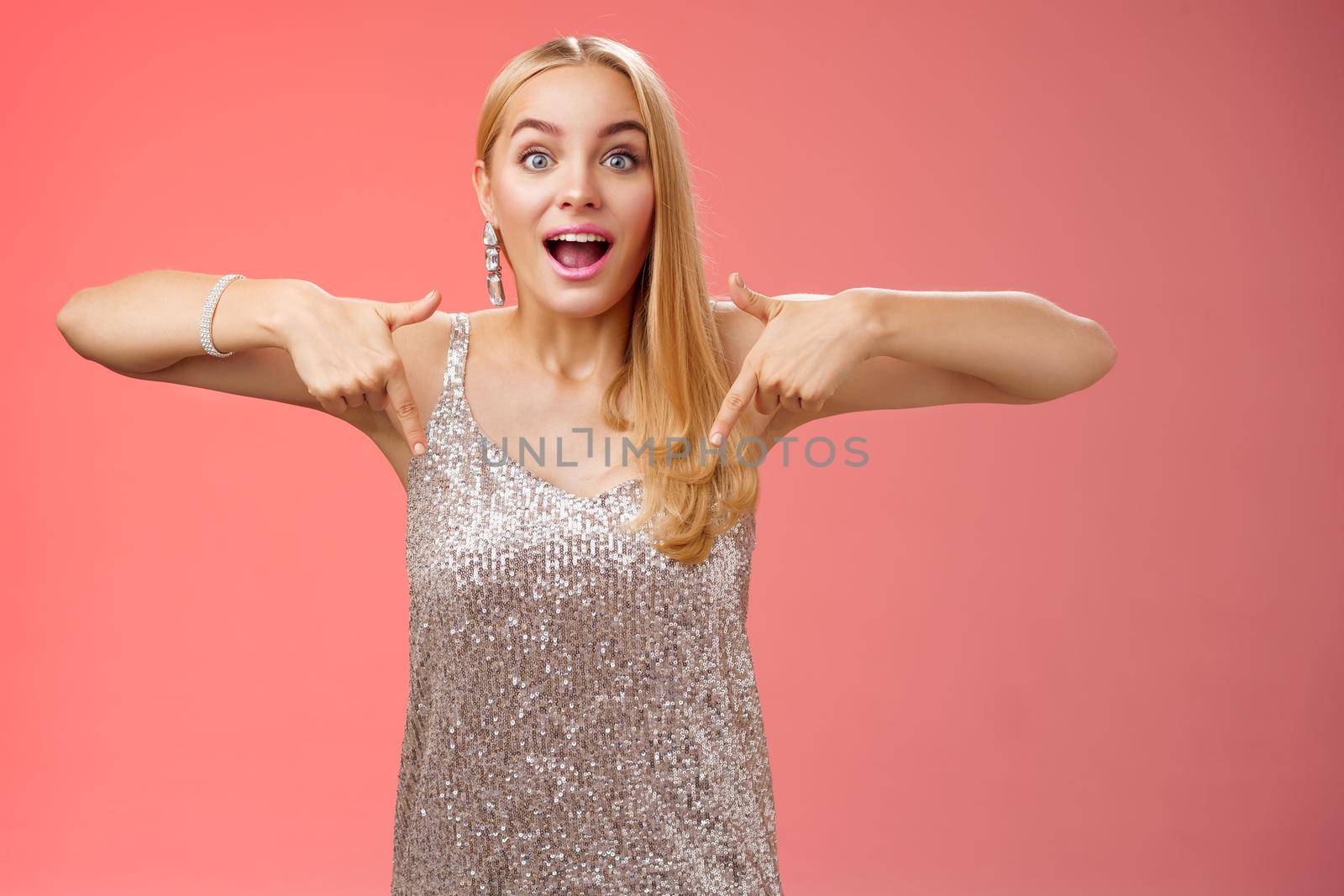 Impressed excited attractive glamour blond girl in silver glittering dress gasping thrilled pointing down glance camera fascinated check out fabulous awesome promo, standing amused red background.