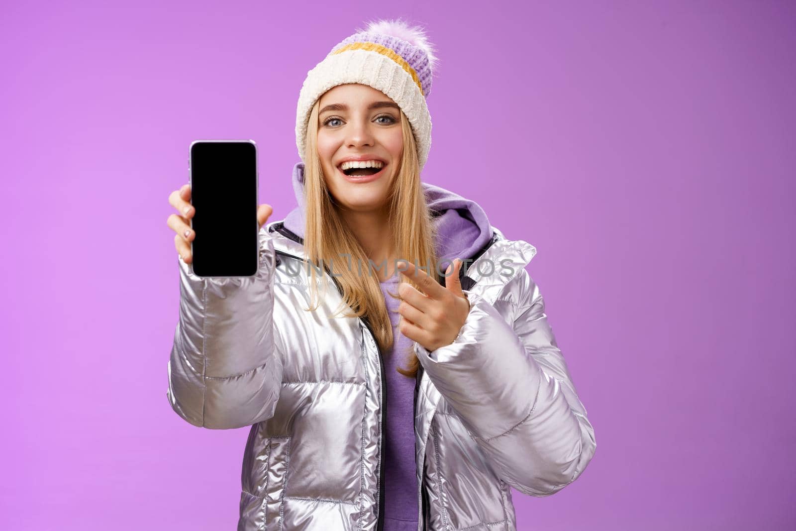 Satisfied amused good-looking blond girl suggest take look smartphone display smiling happily pointing mobile phone delighted talking about awesome new app features, standing purple background.