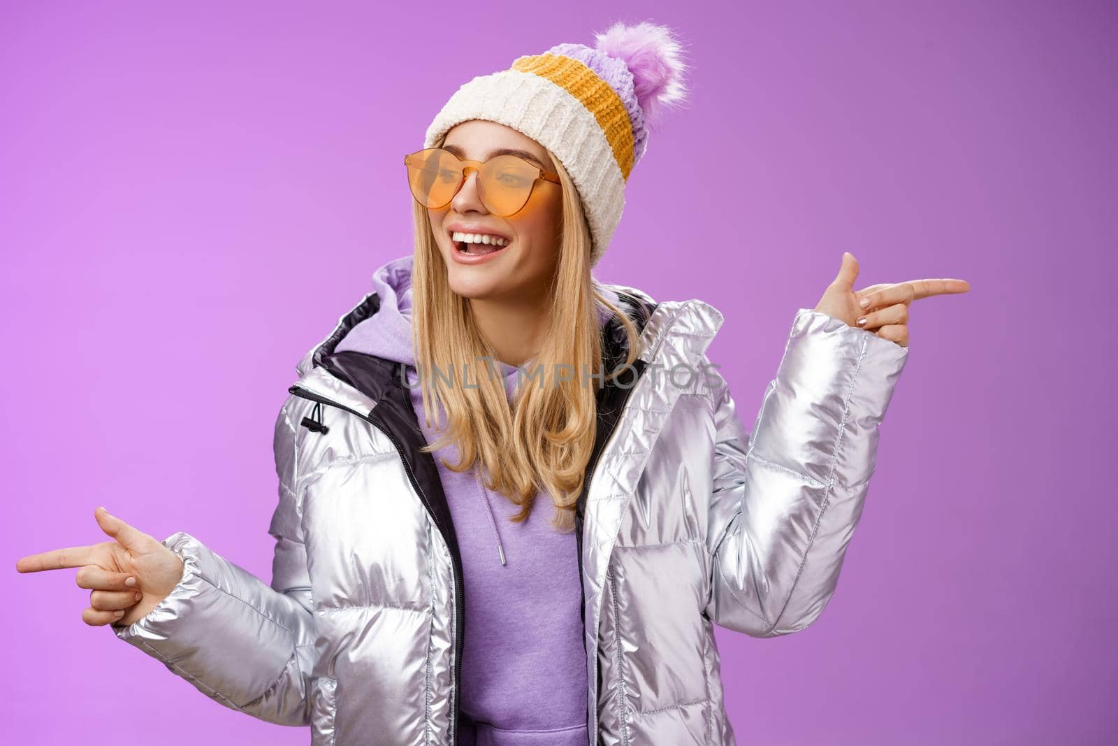 Cool sassy friendly-looking blond girl pointing different sides right left choose which way go look delighted carefree enjoy awesome winter vacation wear stylish silver jacket, hat sunglasses.