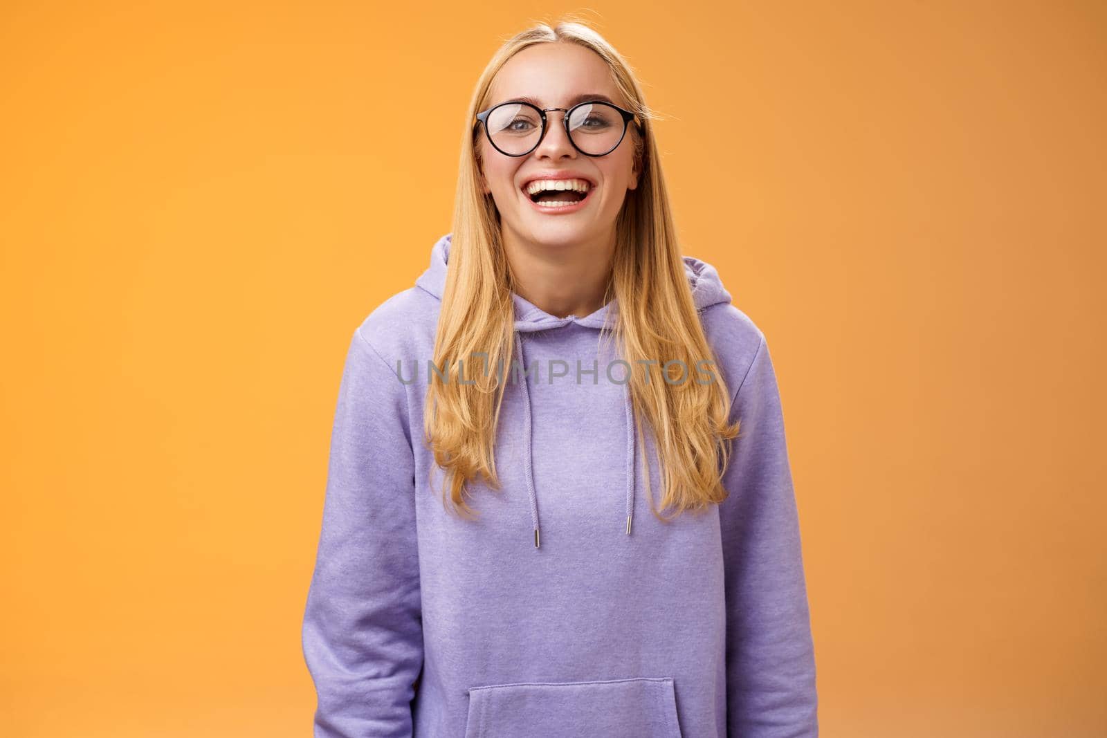 Charismatic joyful charming smiling female university straight a student in glasses purple cozy hoodie grinning laughing happily glad invited hand out classmates standing orange background.