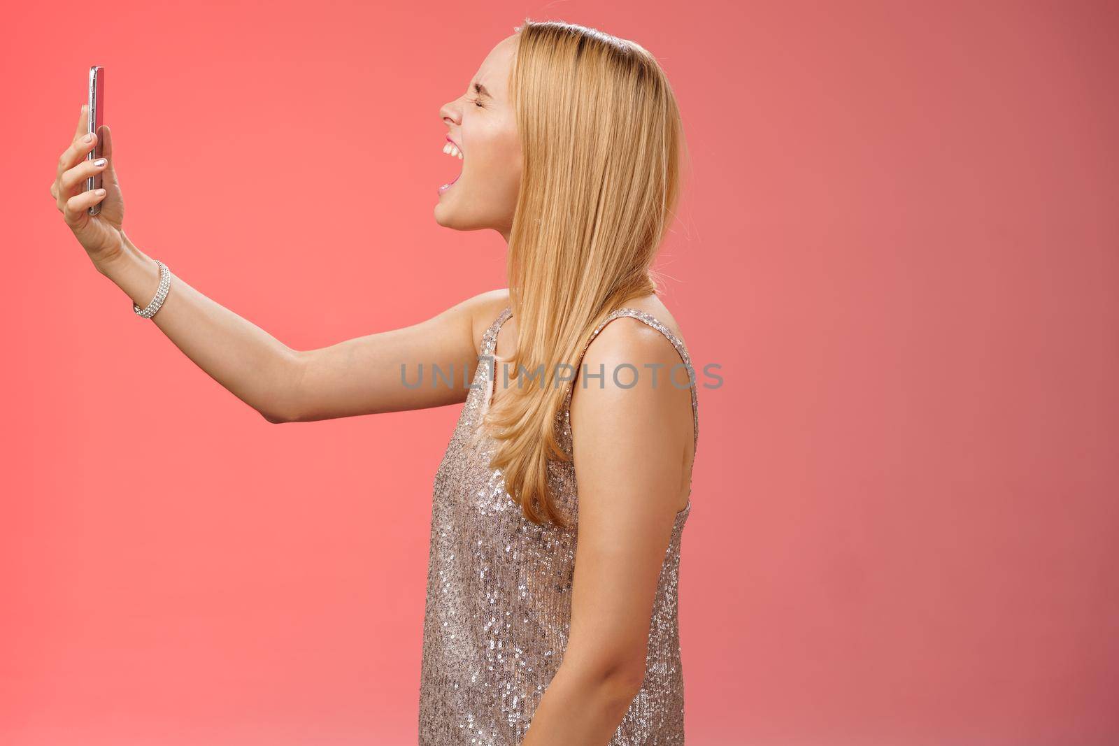 Profile shot funny carefree blond woman hold smartphone raised open mouth wide yelling recording video own shout scream, standing silver glamour dress near red background fool around.
