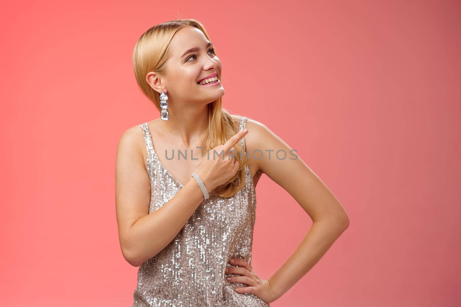 Dreamy tender attractive blond young woman in stylish silver shiny dress pointing looking upper right corner amused smiling broadly delighted see friend peek balcony, standing red background.