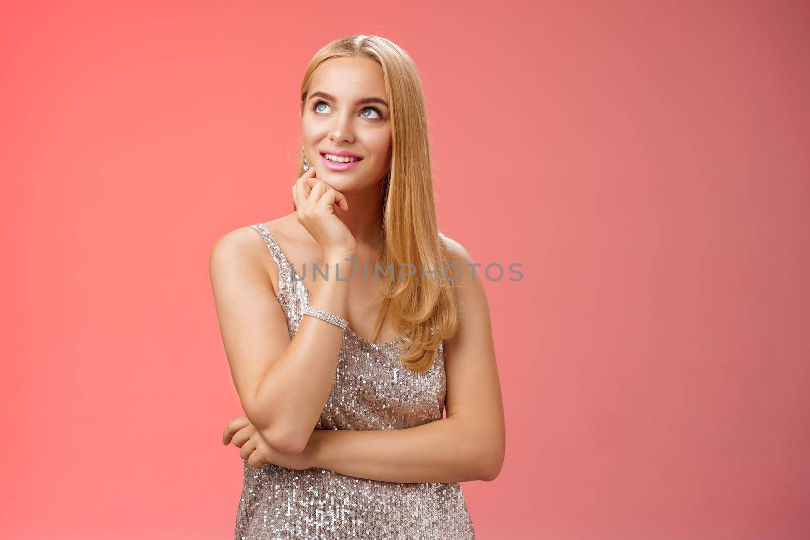 Lifestyle. Dreamy charming elegant blond young woman in silver glittering evening dress look up thoughtful enjoying fashion week afterparty standing casually relaxed red background thinking planning mind.