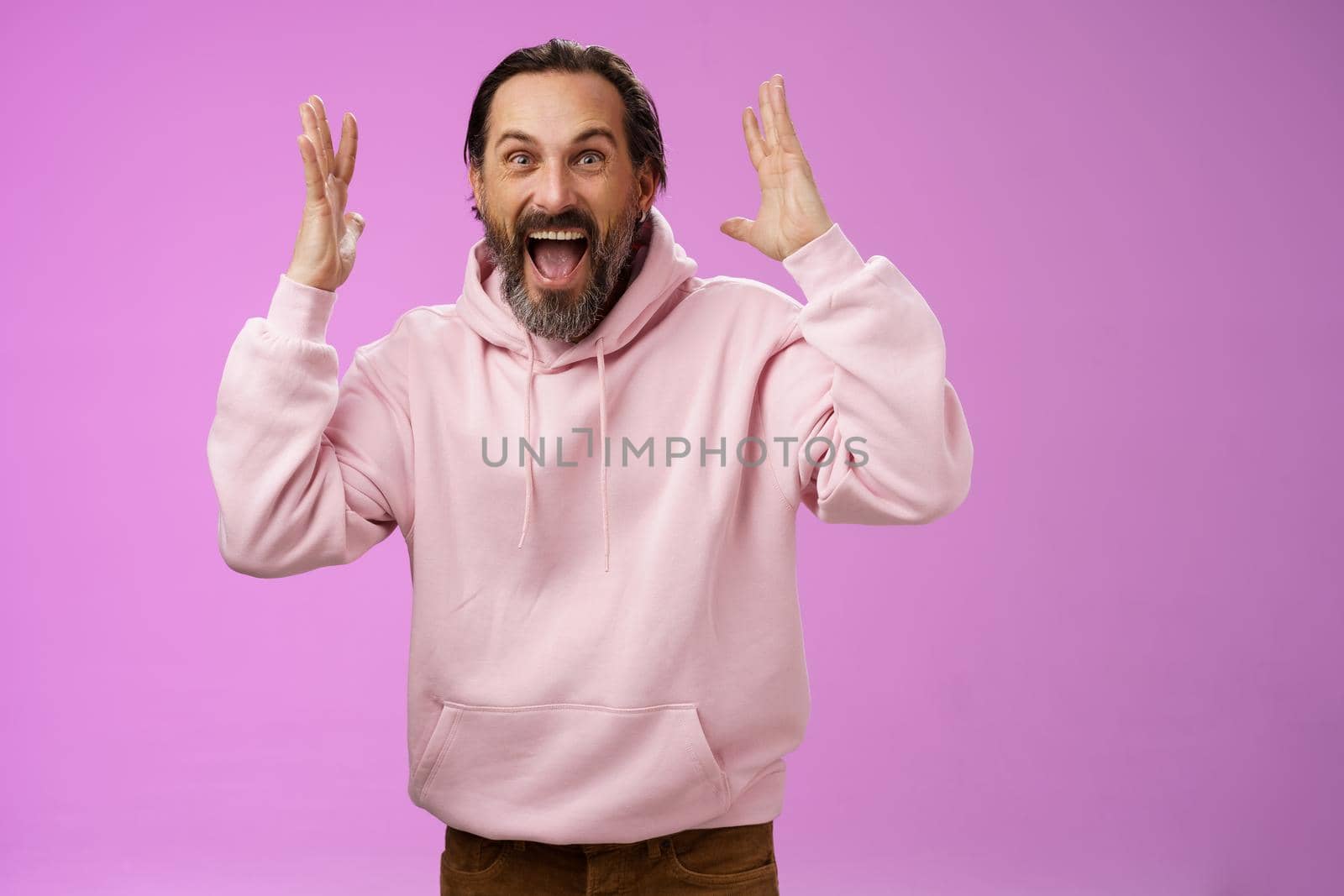 Happy excited lucky adult bearded man celebrating excellent news raising hands gesturing thrilled smiling broaldy triumphing victory win, yelling gladly achieve goal, standing purple background.