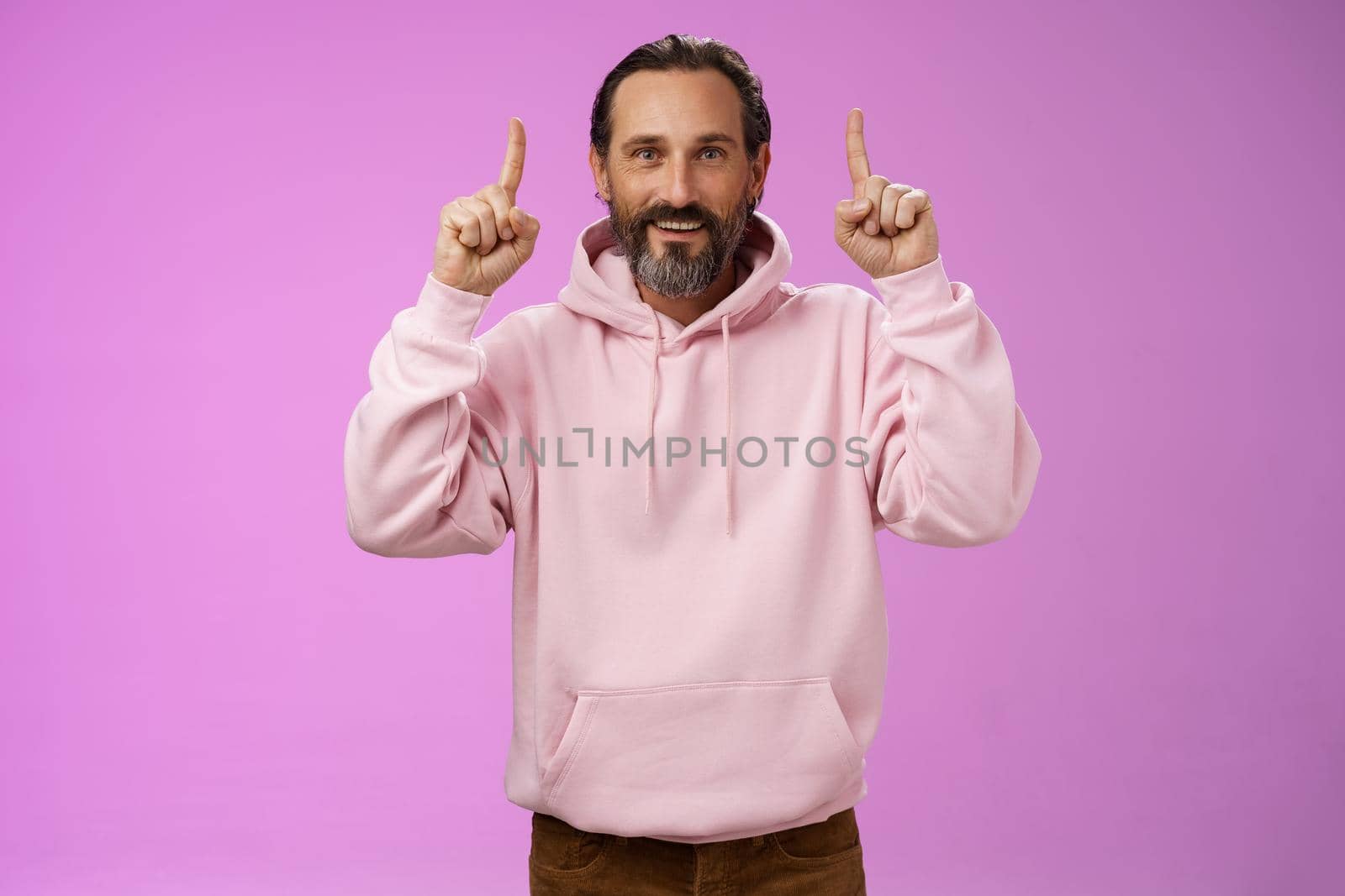 Handsome mature 50s man grey hair beard wearing stylish hipster loose hoodie pointing up index fingers showing awesome place hang out unwilling grow old standing positive lucky purple background. Lifestyle.