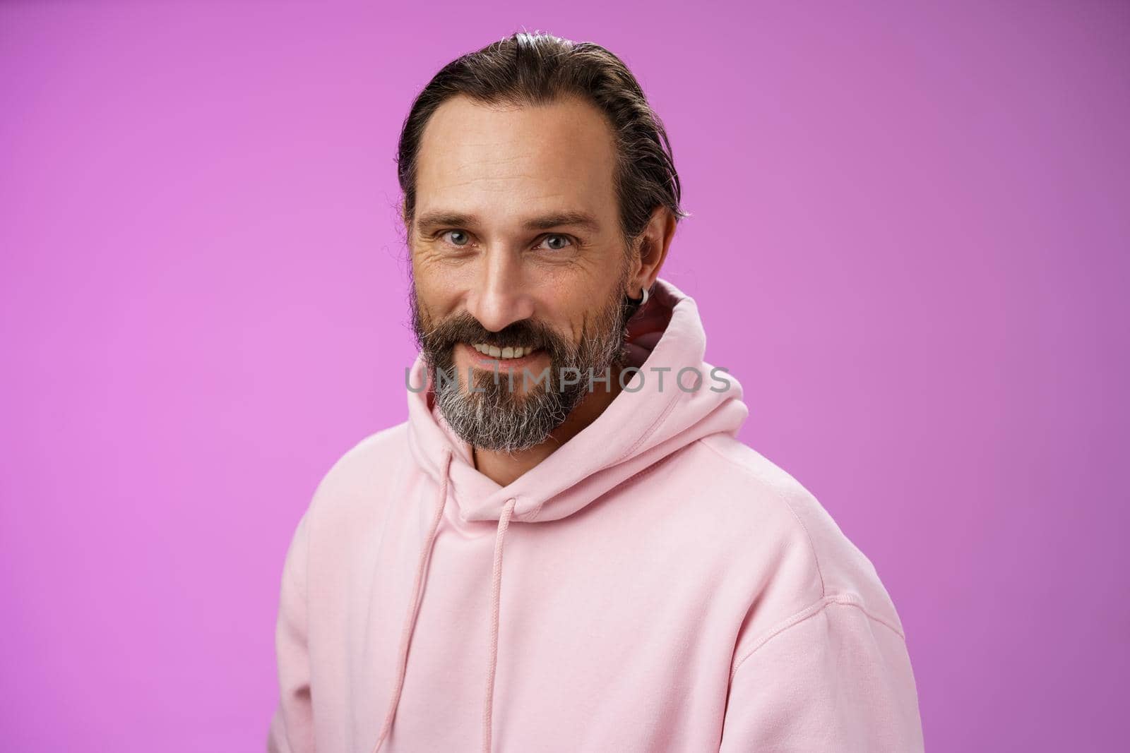 Charming alluring handsome bearded stylish adult male model earring pink hoodie smiling delighted express confidence positivity feel lucky amused, standing purple background talking casually.
