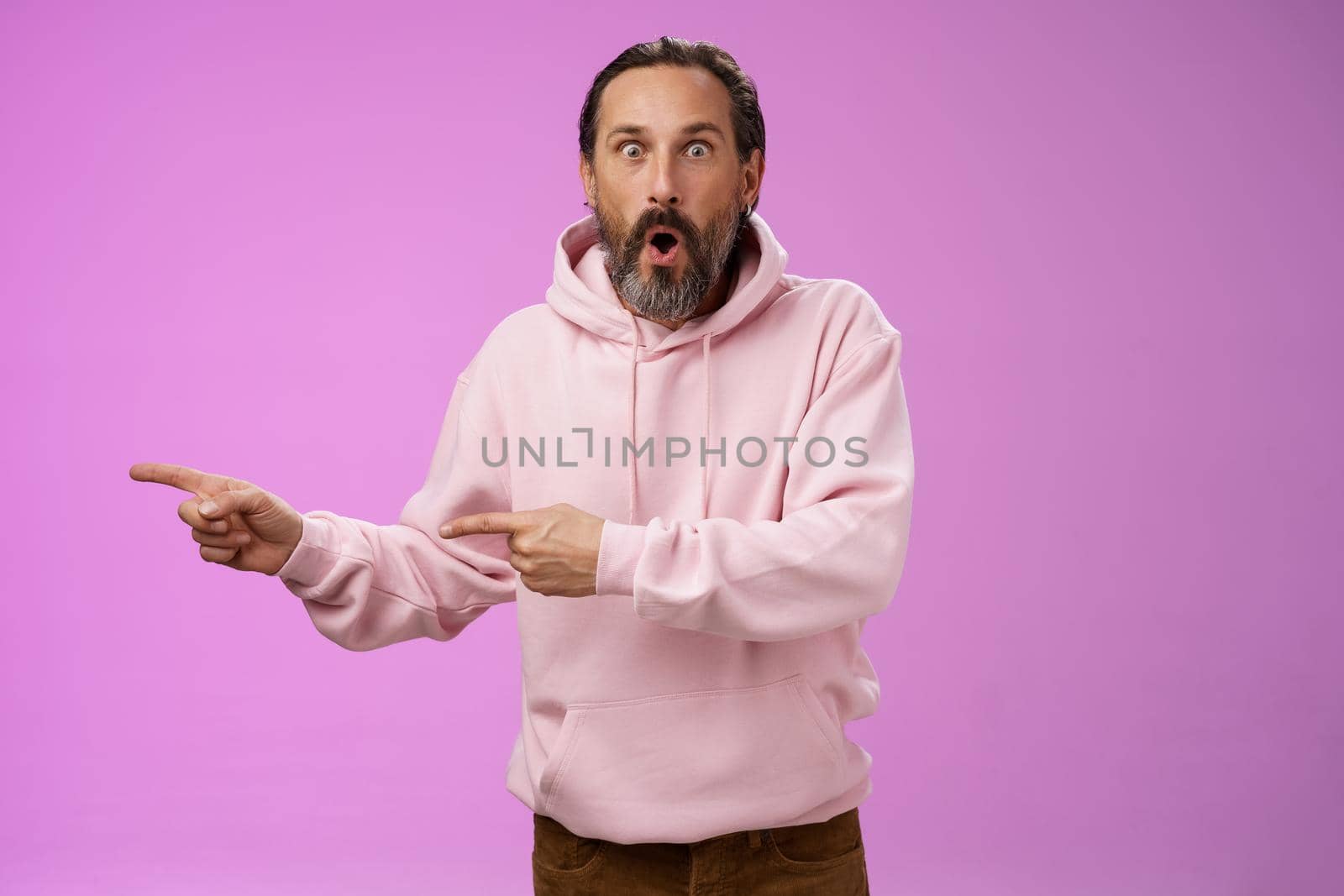 Astonished fascinated curious mature bearded man with grey hair in pink hoodie folding lips amazed pointing left index fingers widen eyes camera impressed thrilled wanna go check out cool promo by Benzoix