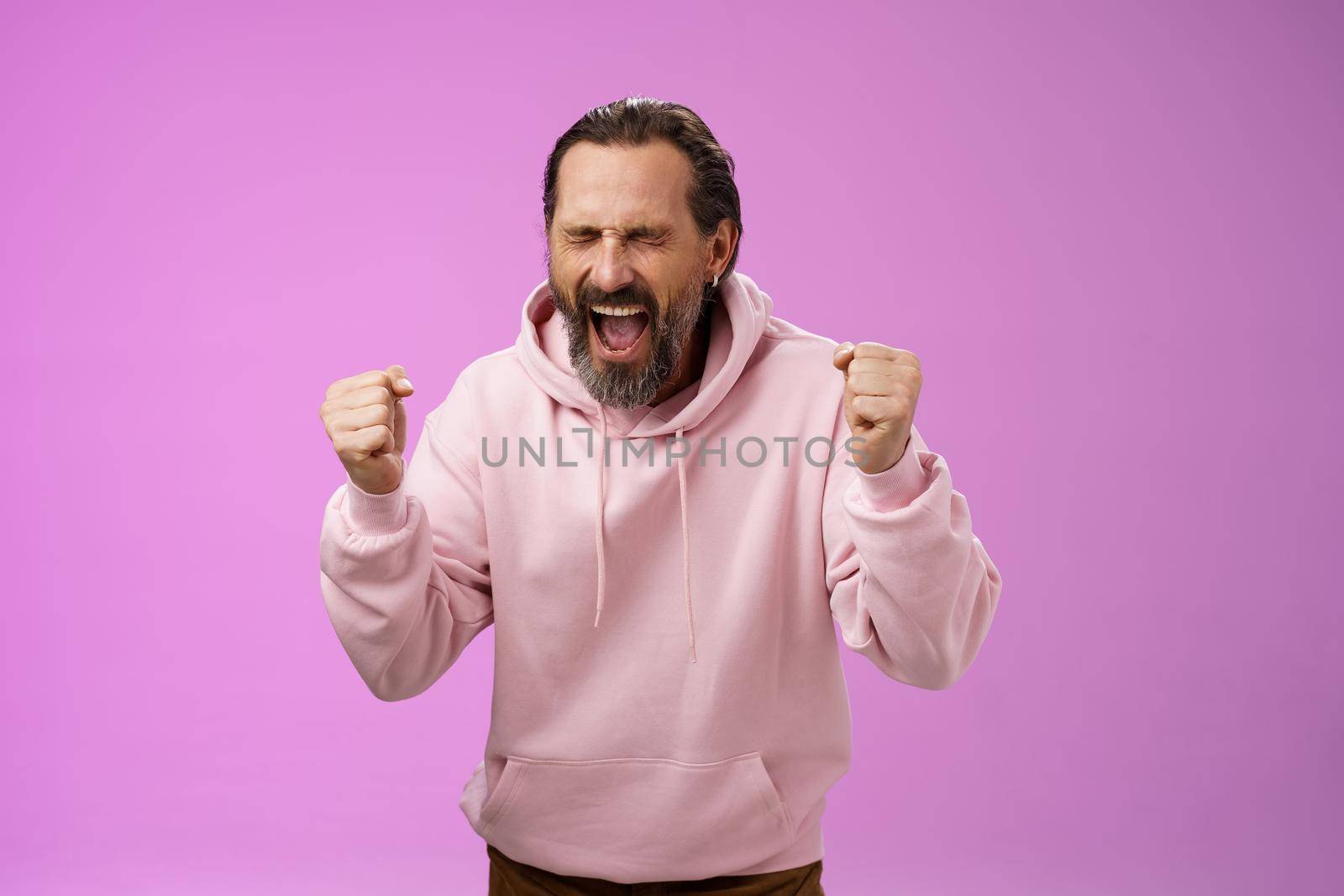 Accomplished happy joyful triumphing mature 40s bearded man grey hair yelling yes thrilled joyfully clench fists celebrating success win lottery become millionaire, standing purple background.