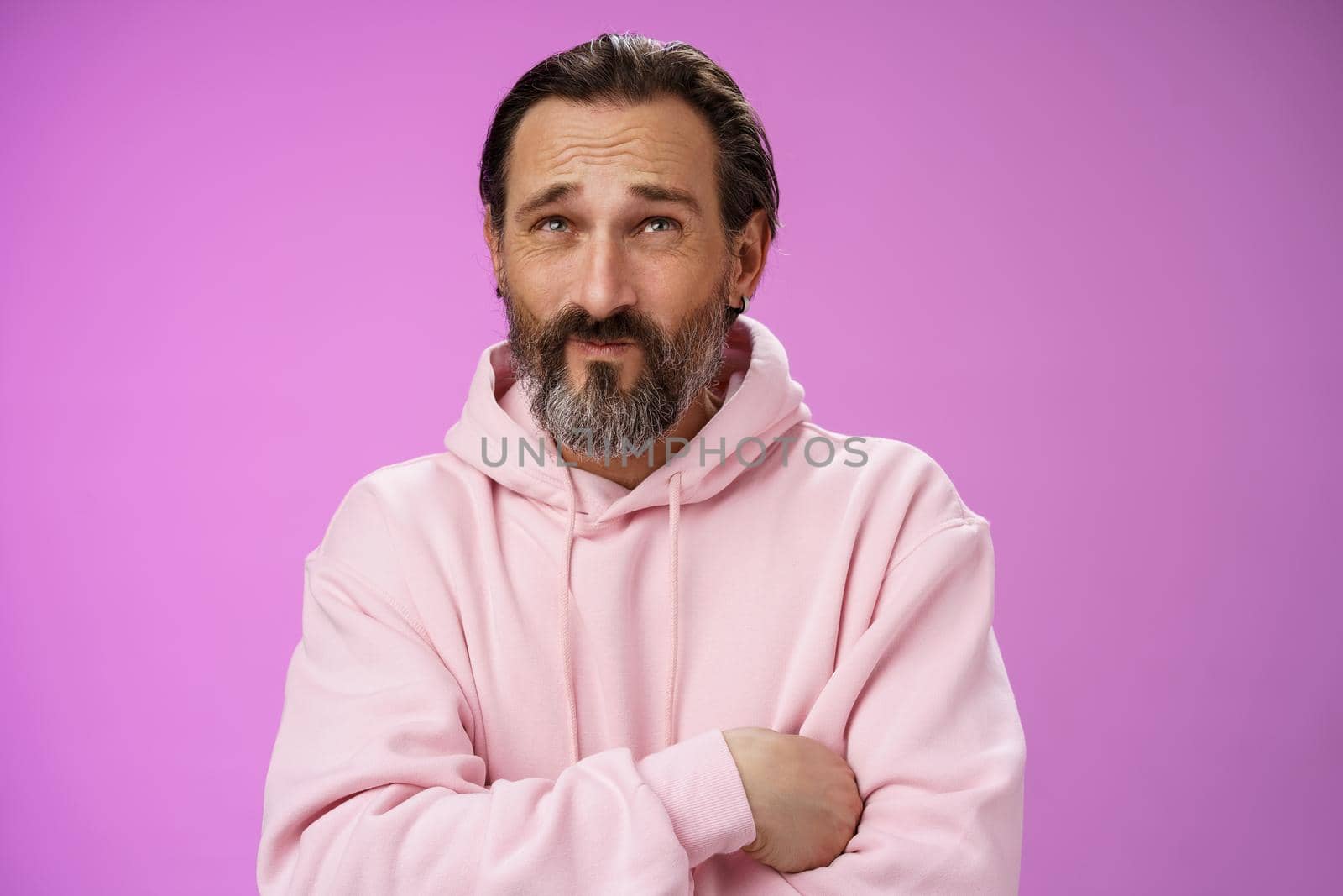 Unsure troubled caucasian bearded adult guy with earring pink hoodie press lips frowning counting mind standing hesitant doubtful thinking look up uncertain taking important decision.