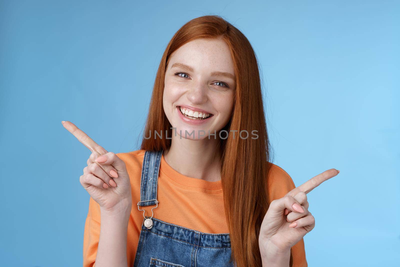 Friendly happy laughing pretty redhead girl female student showing lots opportunities advice make choice pointing sideways left right introducing different product grinning gladly, blue background by Benzoix