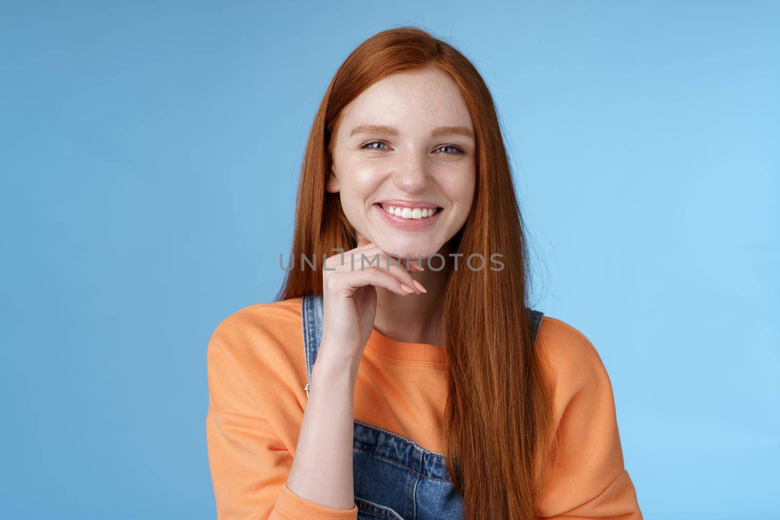 Stylish confident happy smart creative redhead girl look delighted thoughtful like interesting suggestion touch chin smiling approval friendly talking, standing blue background wear overalls by Benzoix