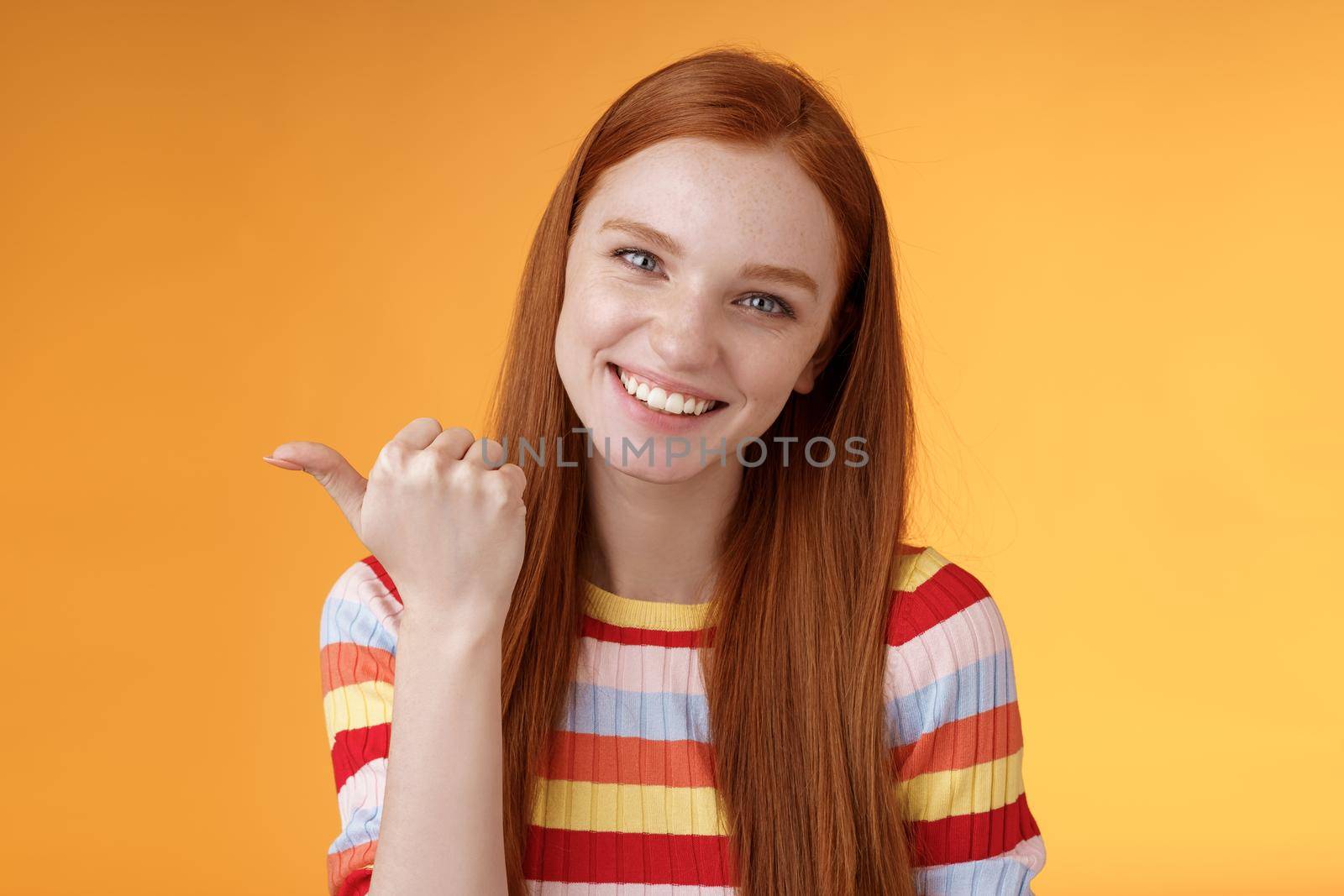 Friendly tender feminine kind redhead woman blue eyes pointing thumb left showing location where find product copy space gladly help discuss interesting promo grinning flirty, orange background.