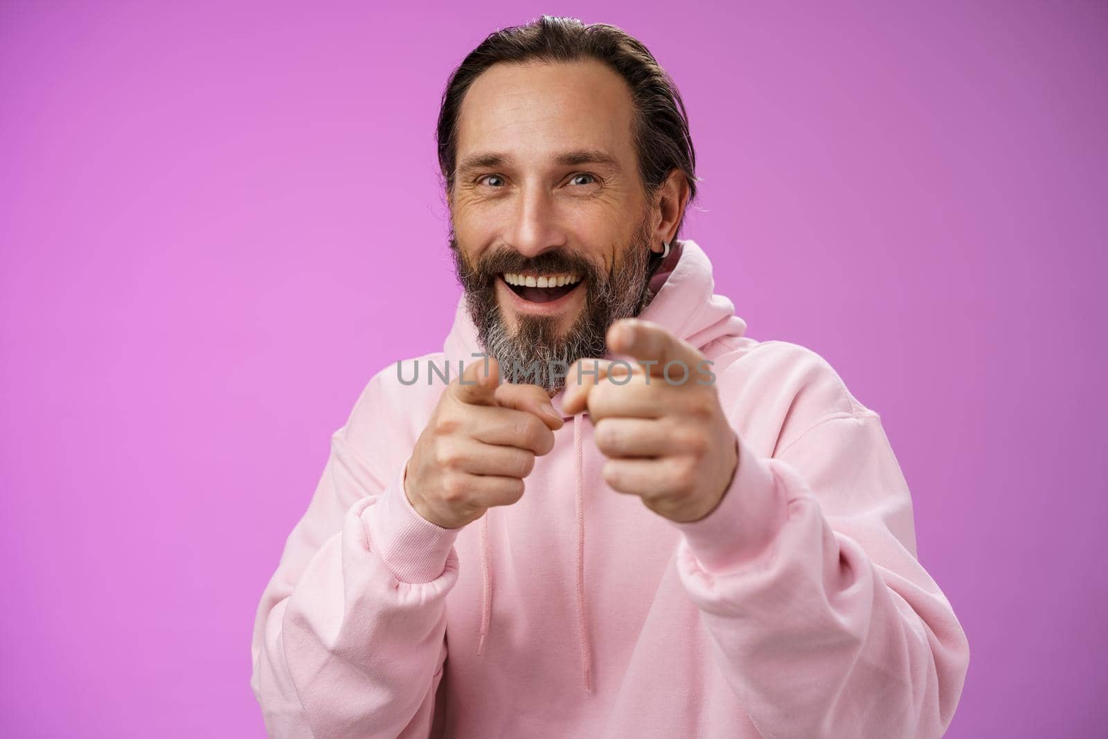 Funny amused carefree happy adult handsome man pranking friend fool around pointing camera index fingers greeting choosing you laughing joyfully having fu, standing purple background rejoicing.