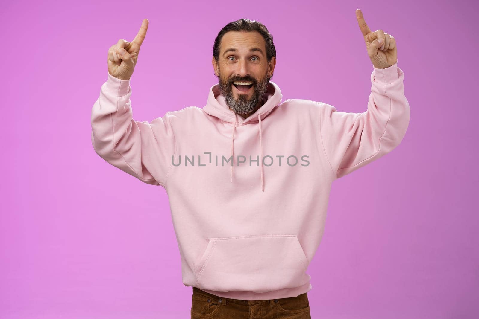 Lucky carefree stylish mature man beard grey hair raising hands joyfully pointing up introduce cool place trying stay young soul wanna hang together grandchildren, standing purple background.