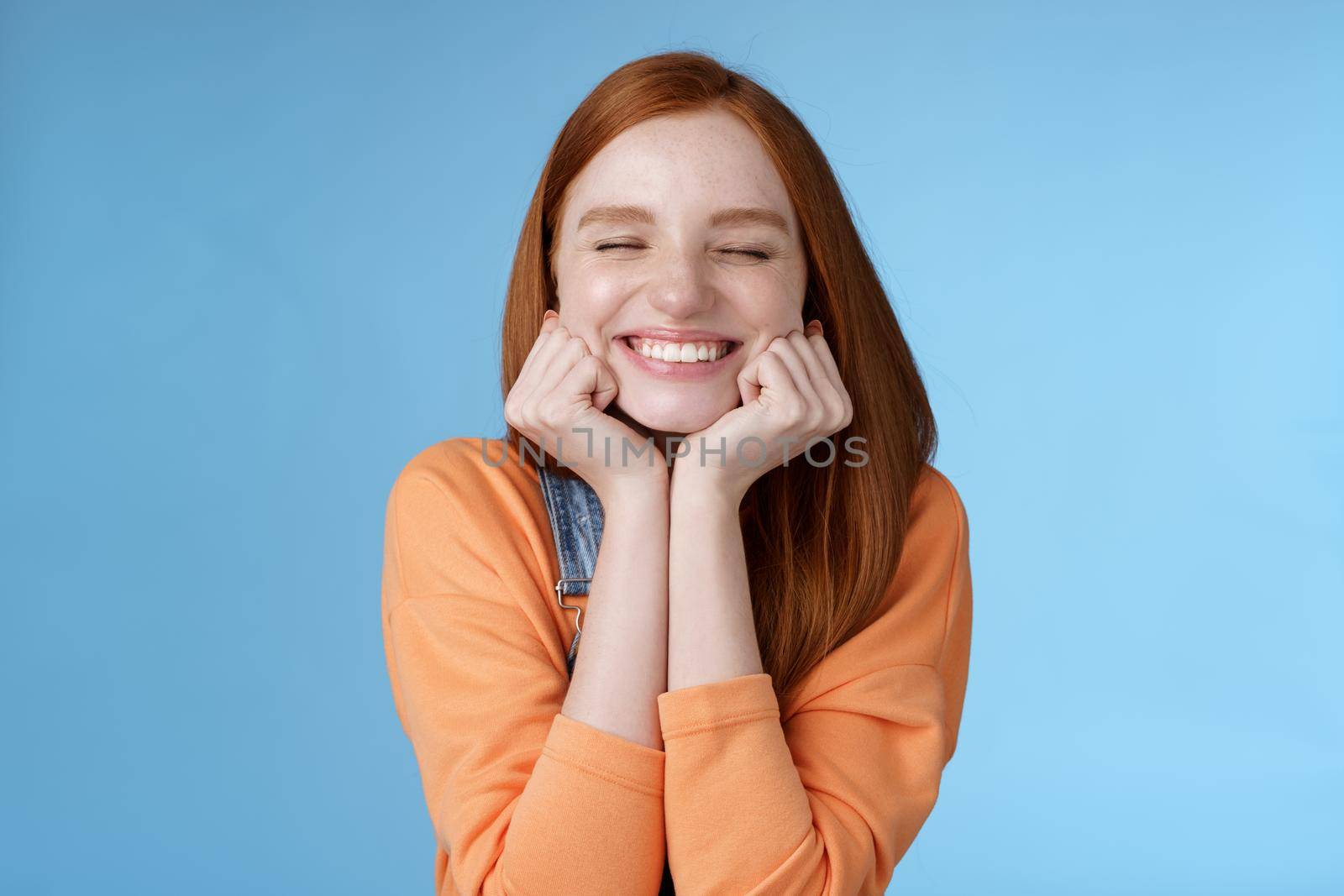 Silly cute happy redhead girlfriend smiling broadly close eyes dreamy squeez cheeks delighted asked date guy likes standing blue background rejoicing have fantastic lucky day triumphing by Benzoix