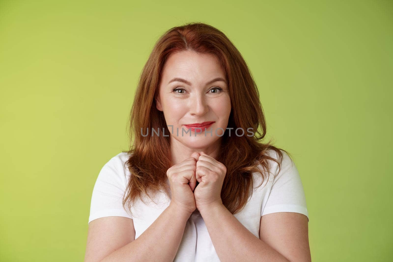 Cheerful lovely pleasant redhead middle-aged female press hands together delight kind happy gaze smiling joyfully touched grateful receive heartwarming gift look admiration happiness.