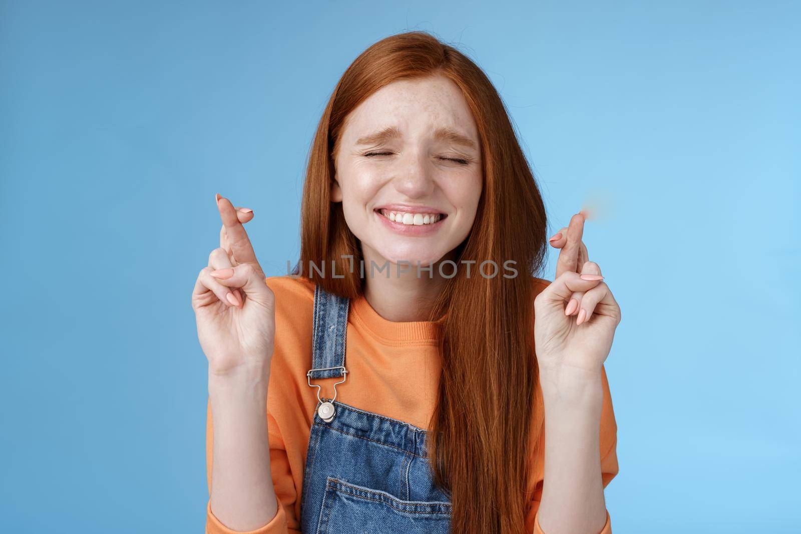 Sincere hopeful cute redhead girl believe miracle close eyes faithfully praying cross fingers good luck hope dream come true make wish eager hear positive results, anticipating blue background.
