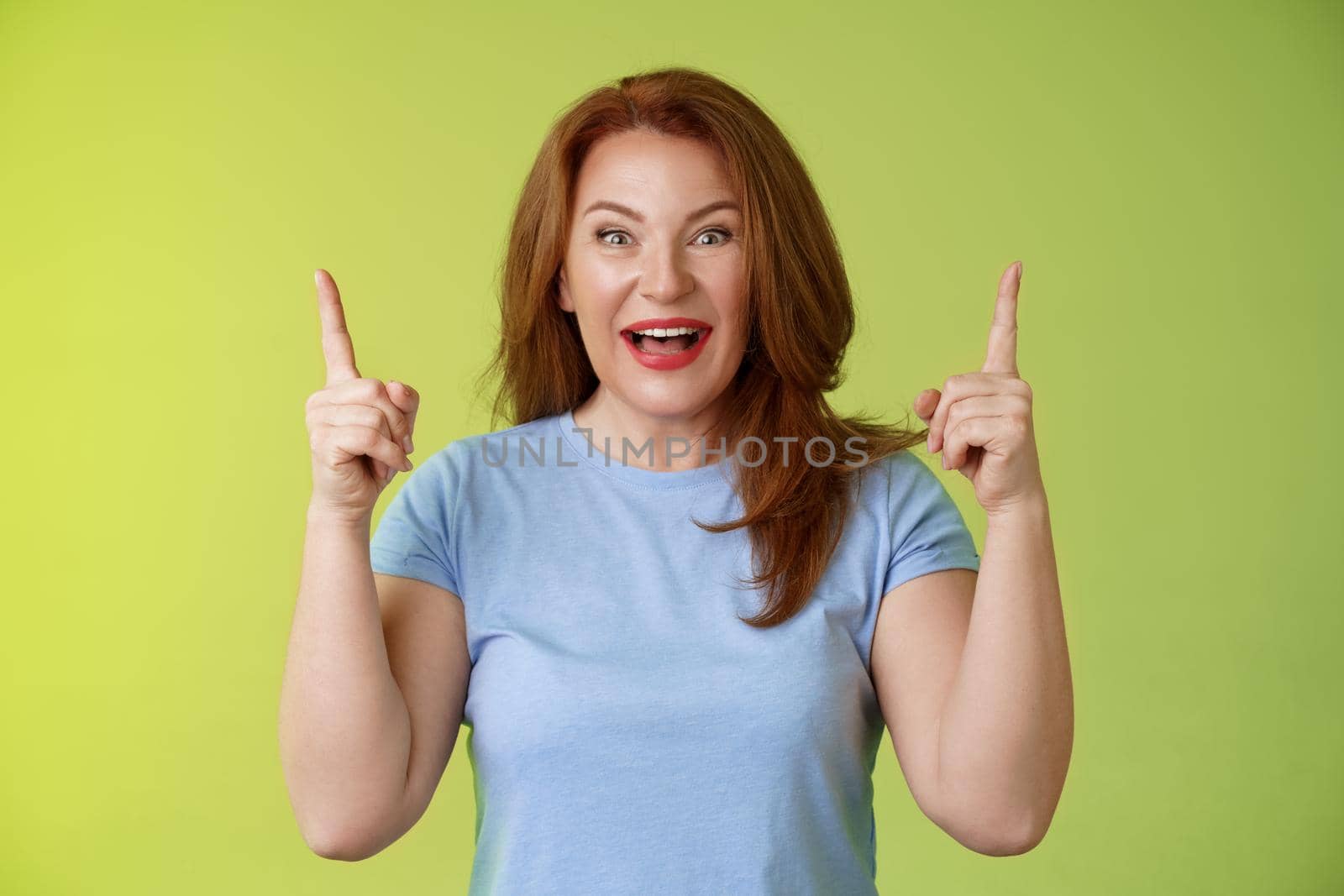 Impressed enthusiastic happy cheerful middle-aged mature woman red hair smiling astonished look admiration joy. pointing up raised index fingers introduce impressive stunning offer green background.