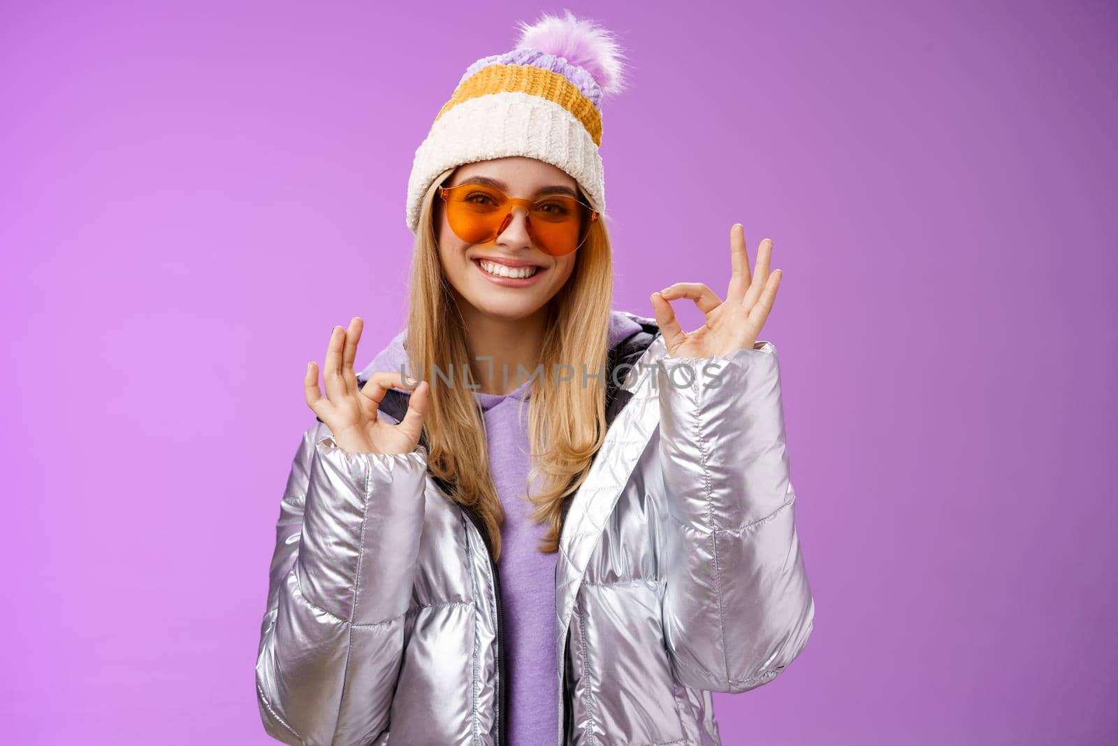 Everything fine thanks. Charming flirty blond confident woman in silver stylish jacket sunglasses winter hat show okay no problem ok gesture smiling affirmative, liking awesome day purple background.