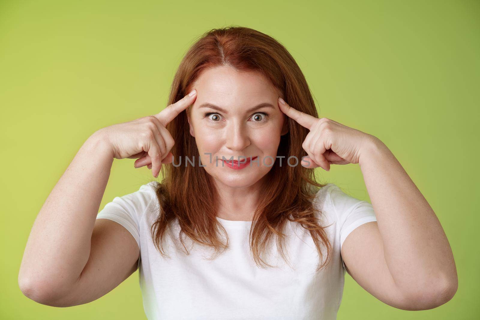 Woman playfully staring funny control your mind. Silly redhead middle-aged female touch temples popping eyes smiling delighted read thoughts trying guess intrigued what thinking green background by Benzoix
