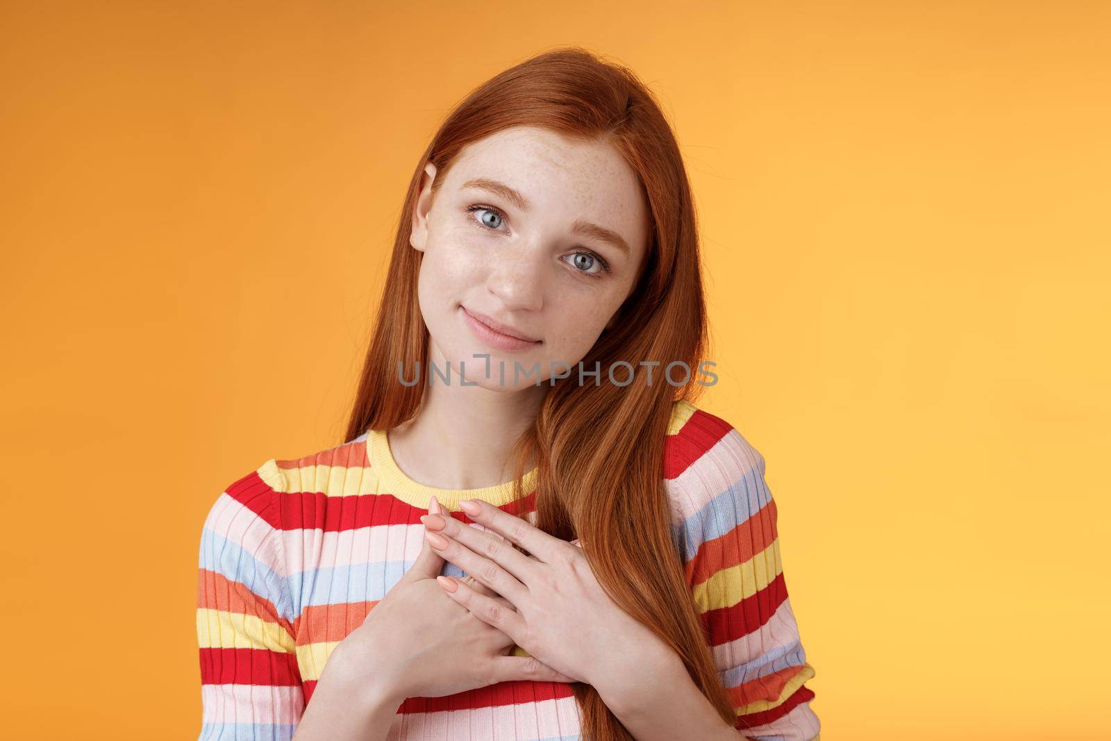 Lovely silly cute redhead young european girl tilting head look gently smiling tender camera press palms heart thankful receive nice gift gazing gratitude happiness, standing orange background.